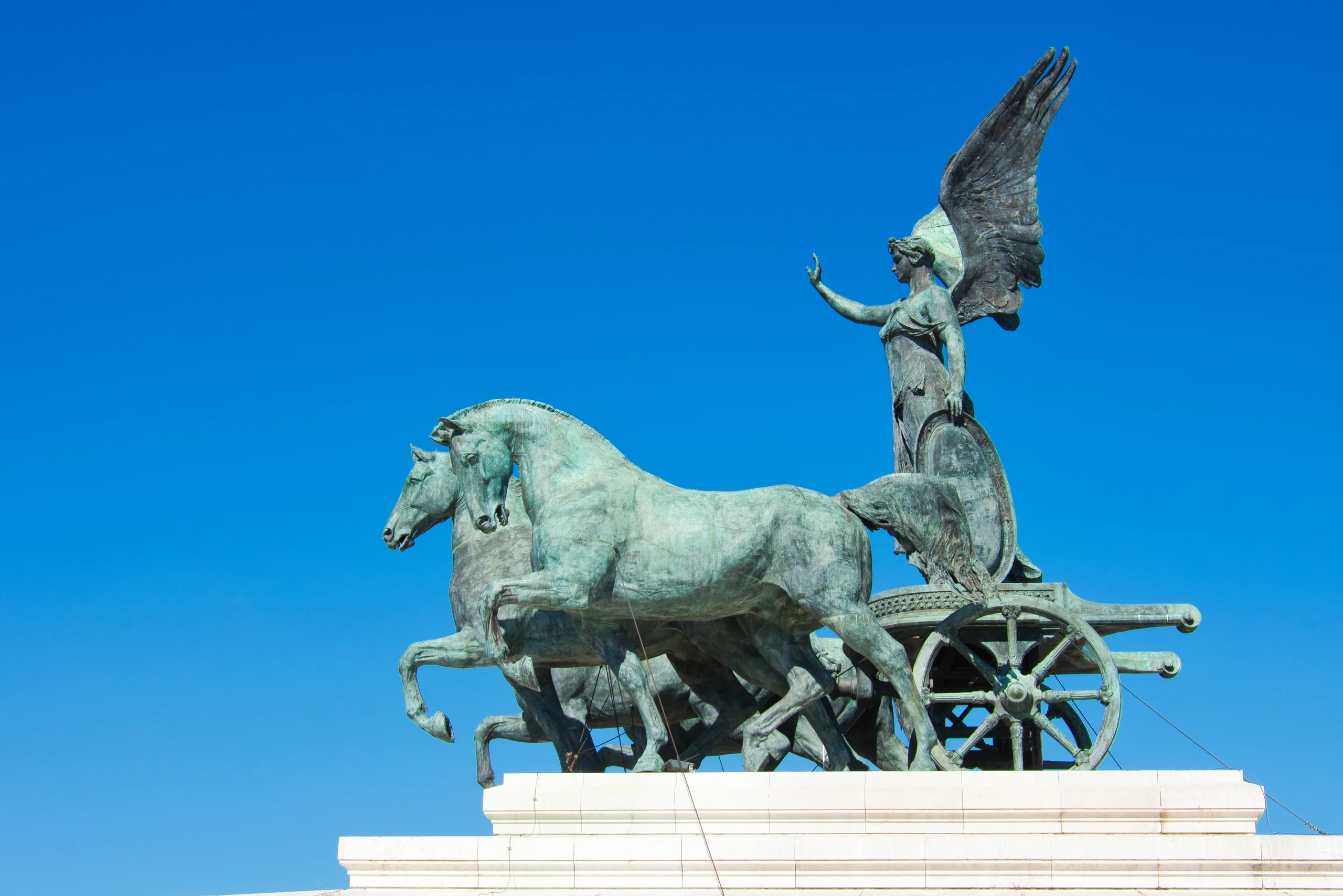 A bronze quadriga on the roof of the Vittoriano, winged Victory driving four horses against an empty blue sky.