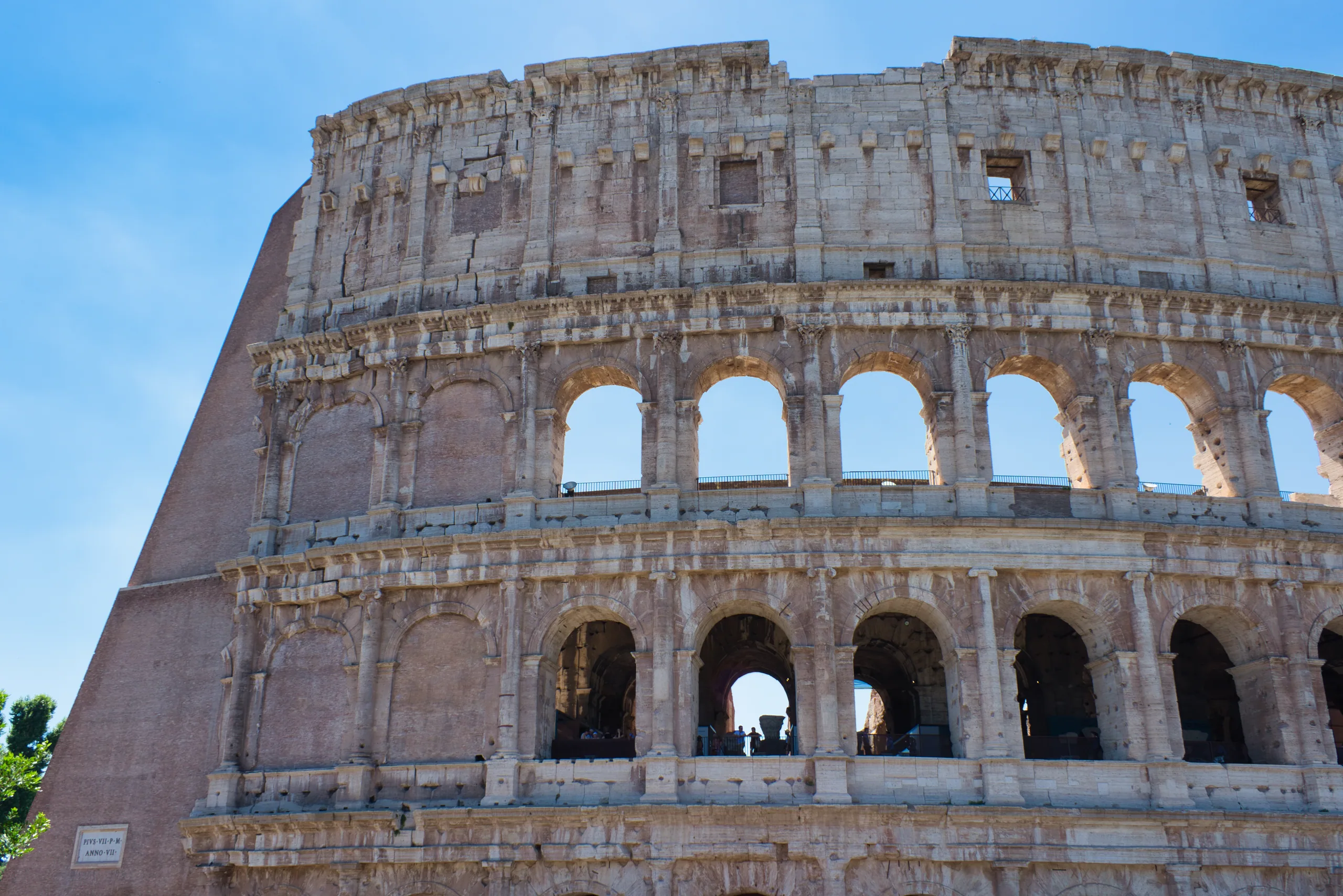 The outer wall of the Colosseum, two tiers of arches curving away against a clear blue sky.
