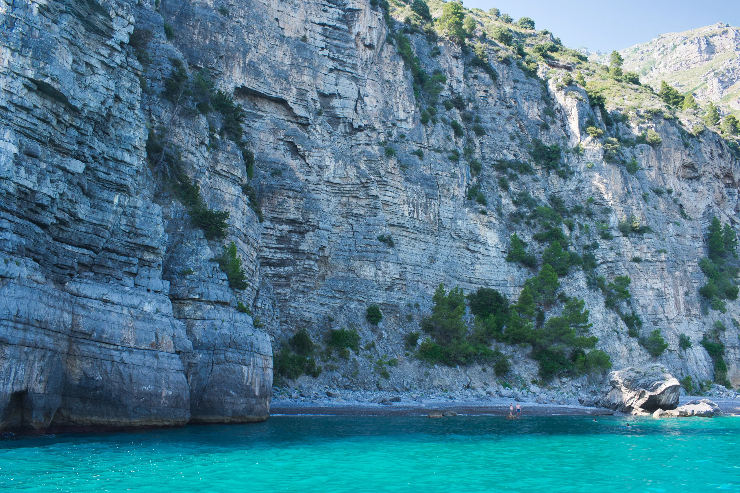 A limestone cliff dropping straight into brilliant turquoise water along the Amalfi coast, swimmers in the shallows below.