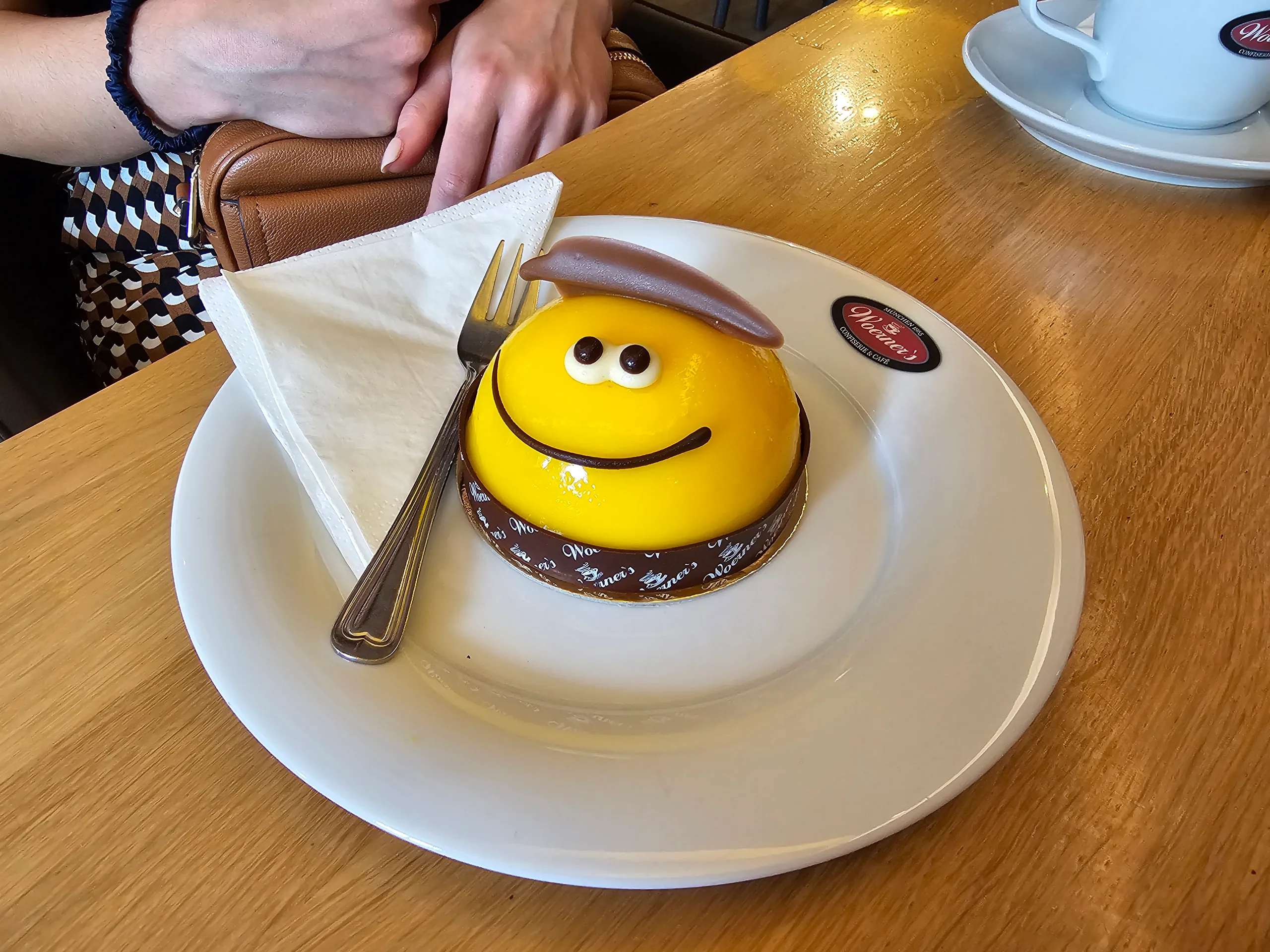 A yellow domed cake decorated as a smiling bee on a cafe plate, a coffee cup beside it.