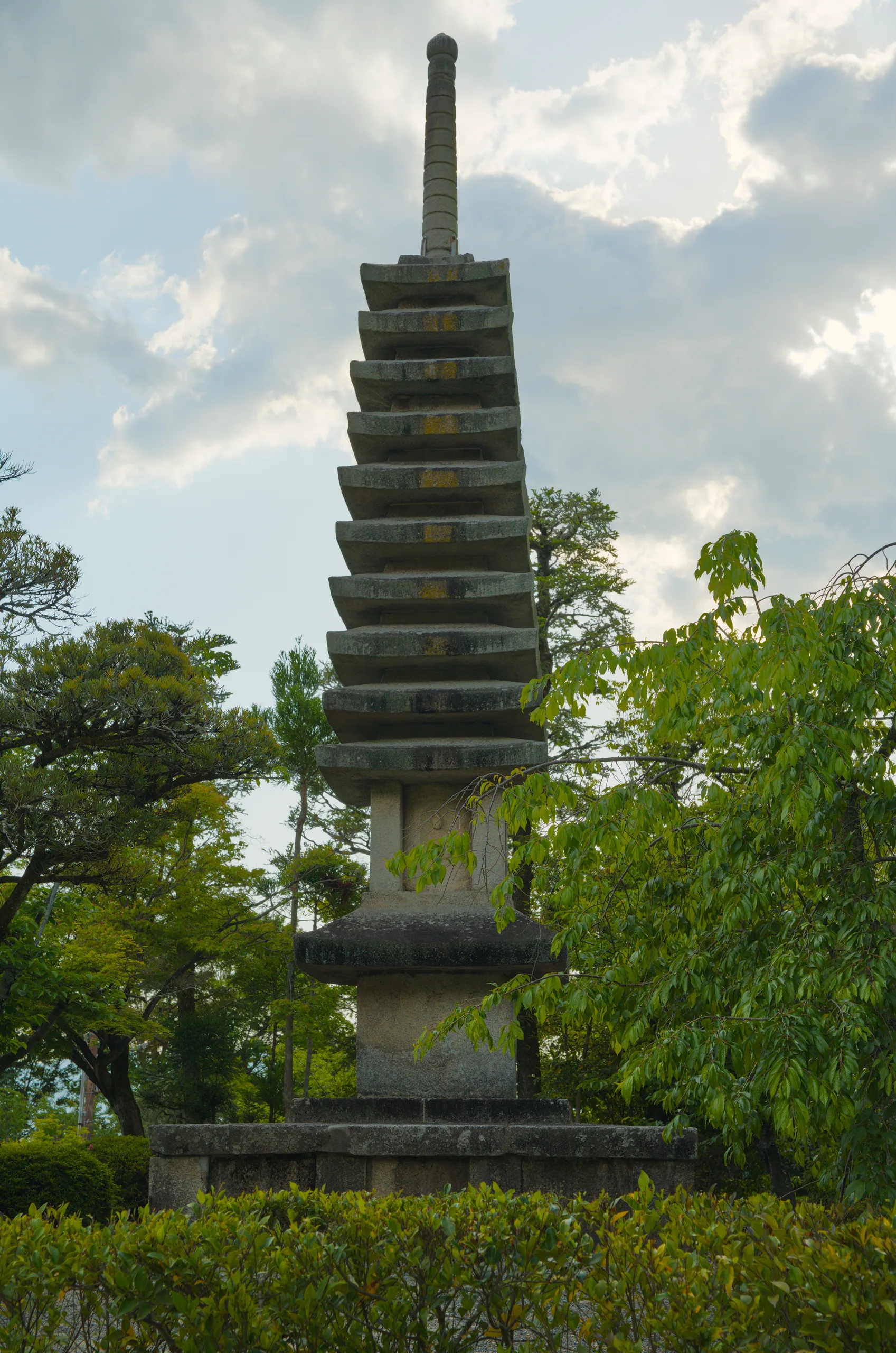 Tall stone pagoda with stacked tiers rises above surrounding trees beneath cloudy sky.