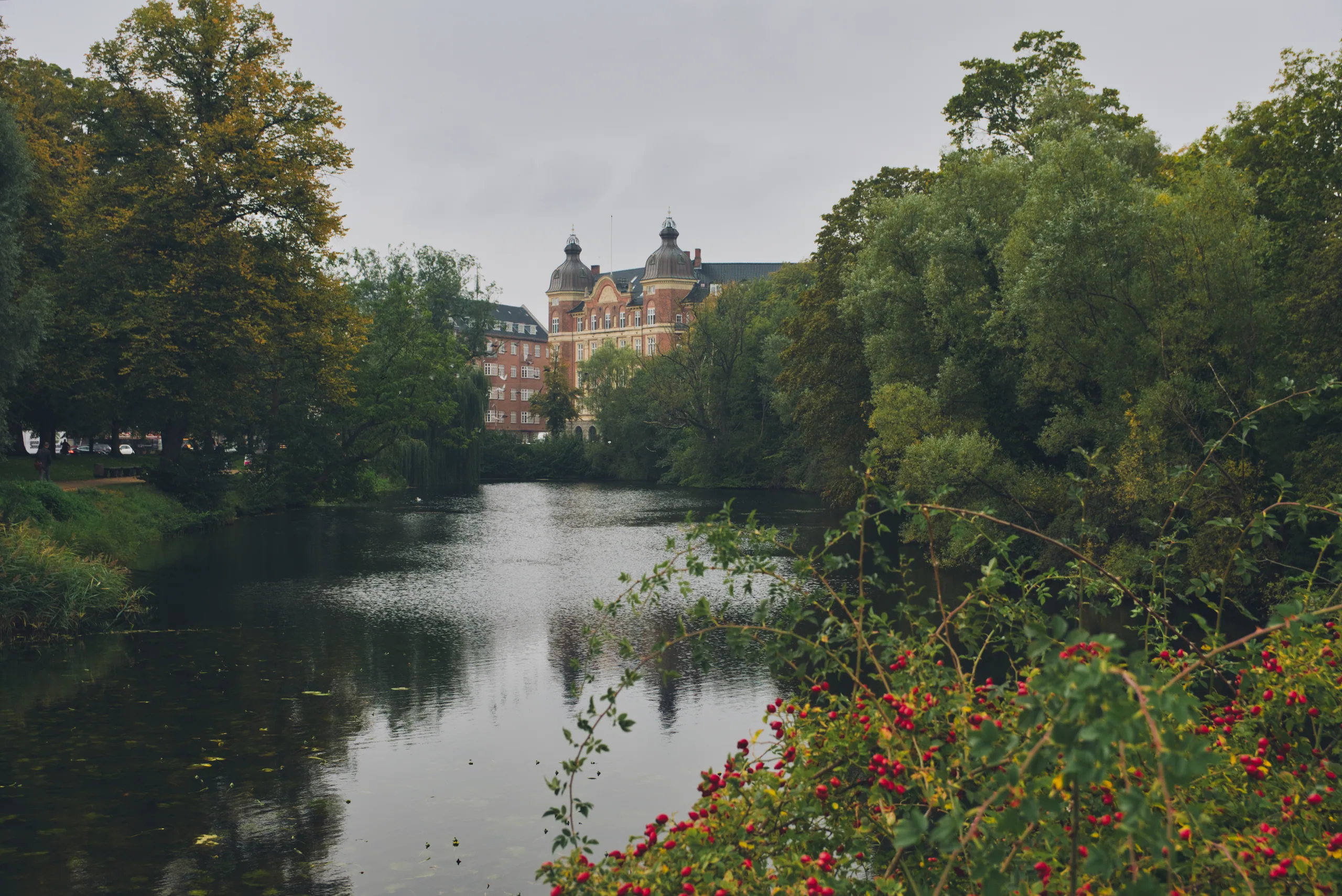 A still canal reflecting trees, a red-brick mansion visible through the foliage and rosehips along the near bank.