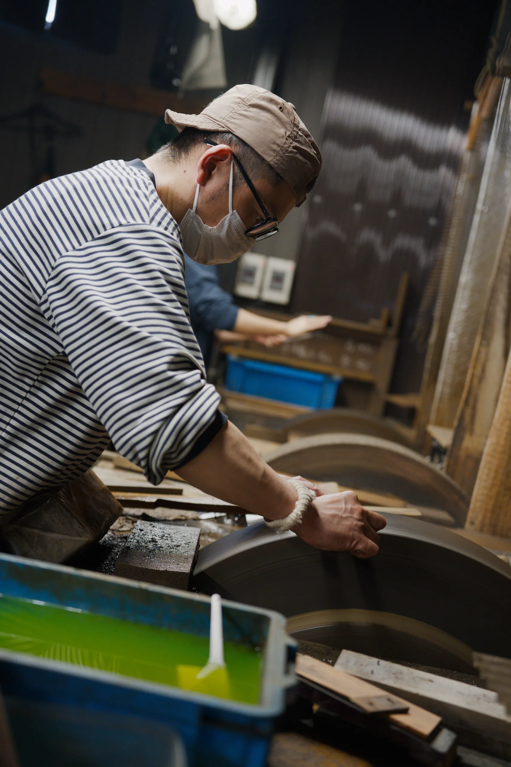 A person in a striped shirt and cap shapes a wooden curved piece in a workshop.