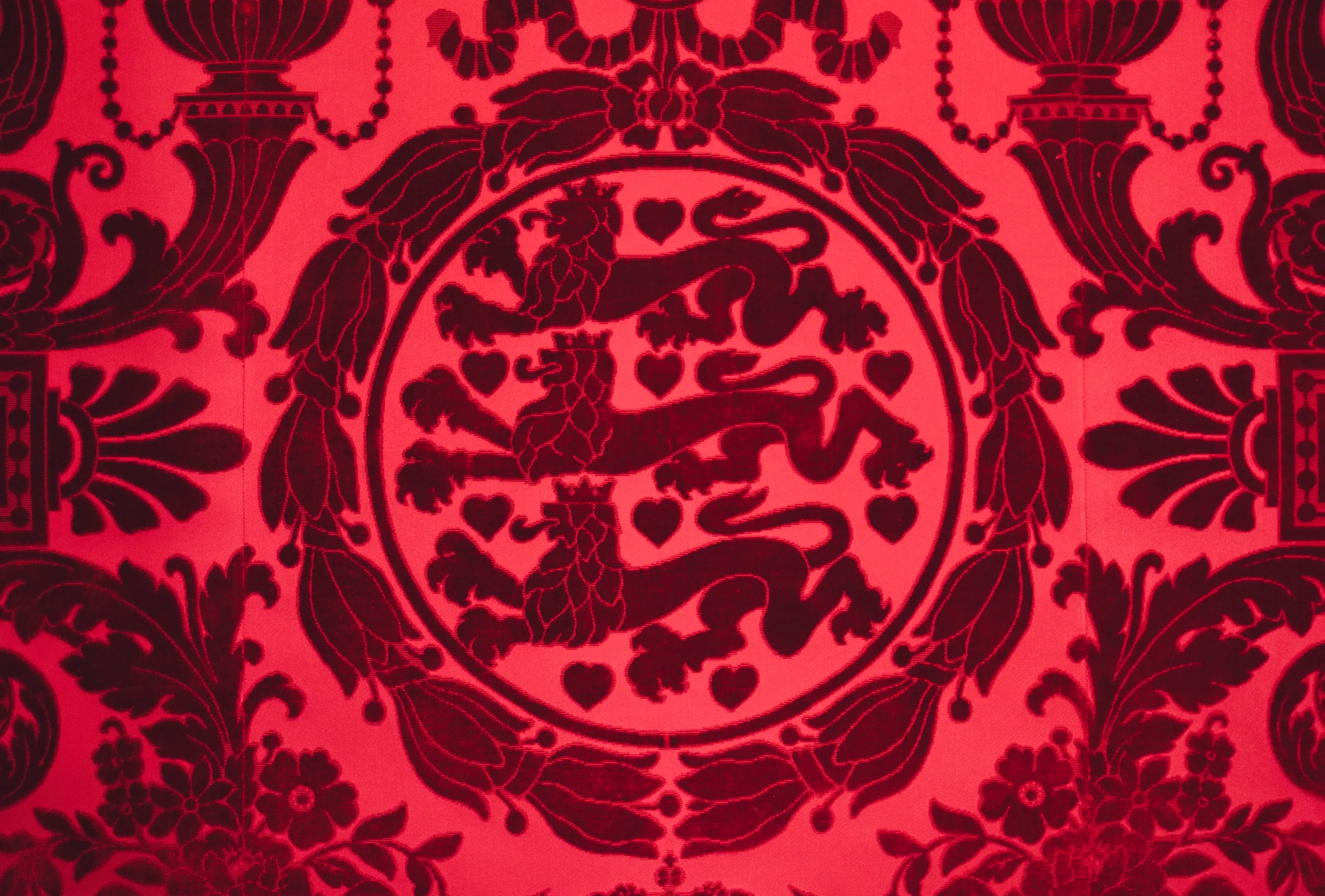 Red damask fabric printed with the Danish coat of arms, three lions and hearts inside a scrolled medallion.
