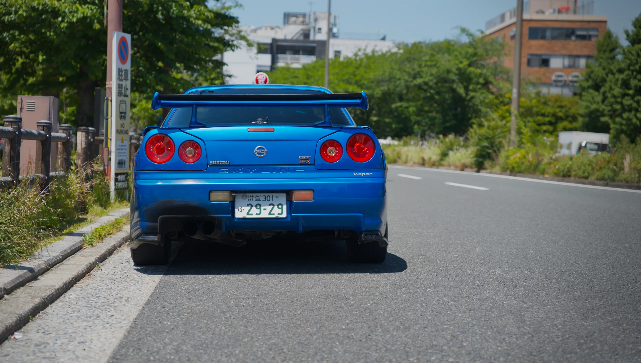A blue Nissan Skyline sports car viewed from behind on a sunny road.