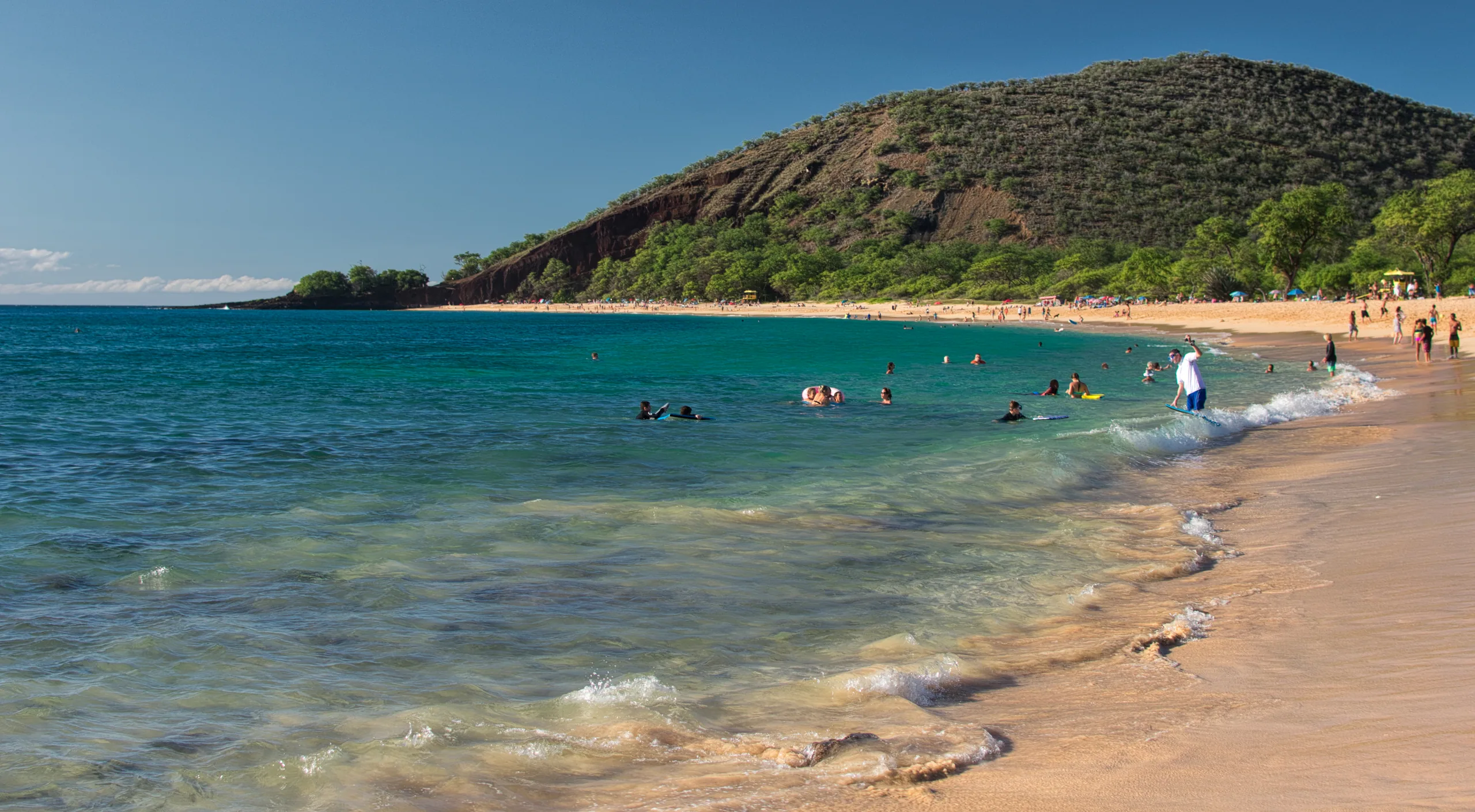 Clear shallow water washing onto a broad sandy beach, swimmers in the shallows and a green cinder cone behind.