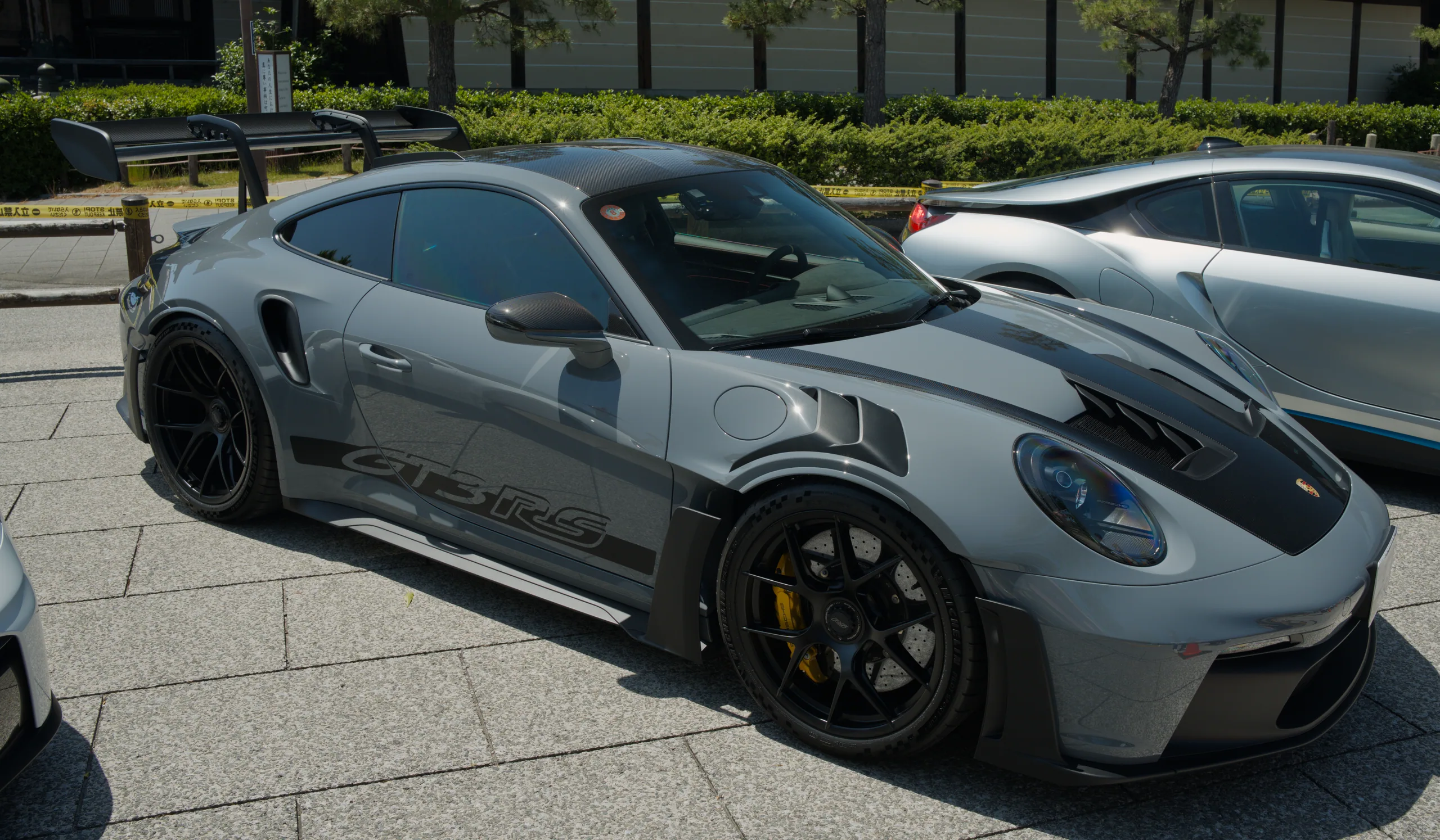 A gray Porsche 911 sports car with red-tiled roofed buildings and green trees in the background.