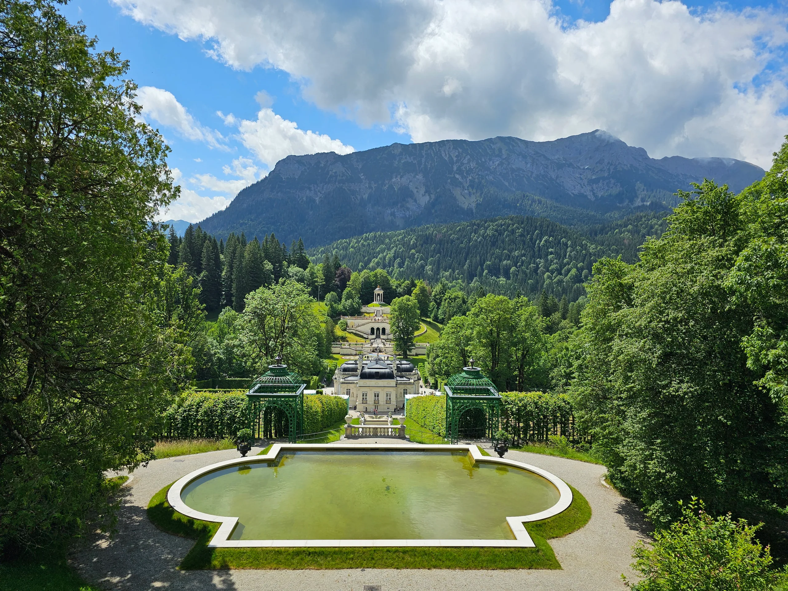 The gardens of Linderhof from above, the oval basin and terraced parterres running up to a wooded ridge and grey mountains.