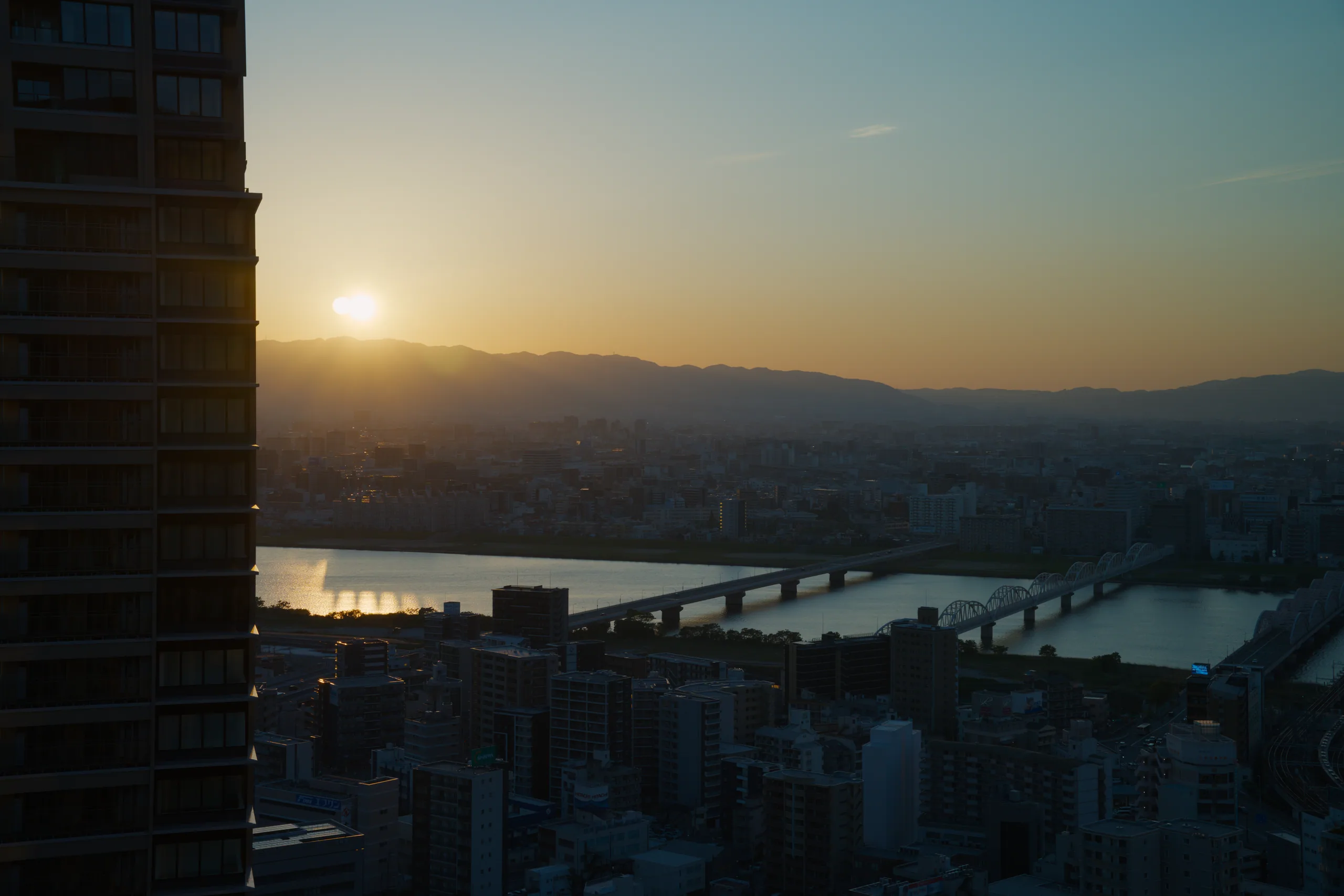 River with multiple bridges crossing through a valley surrounded by mountains and city sprawl at sunset.
