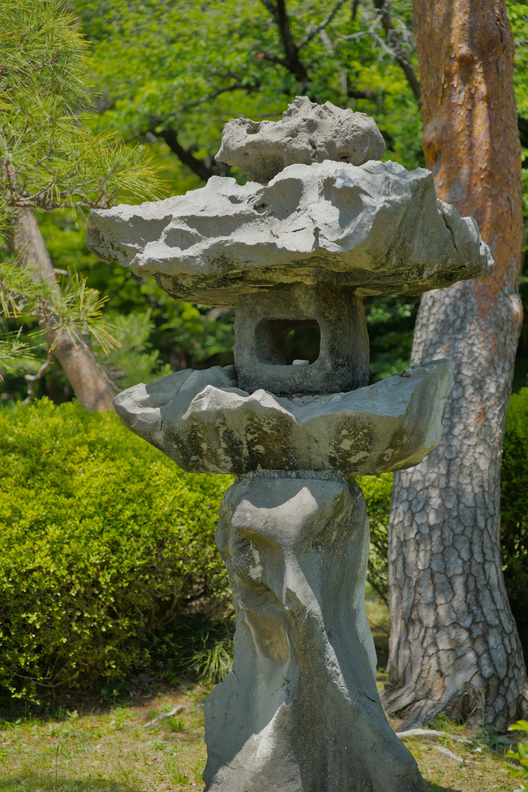 A stone lantern rests on moss-covered ground beside tree trunks surrounded by ferns in a garden.