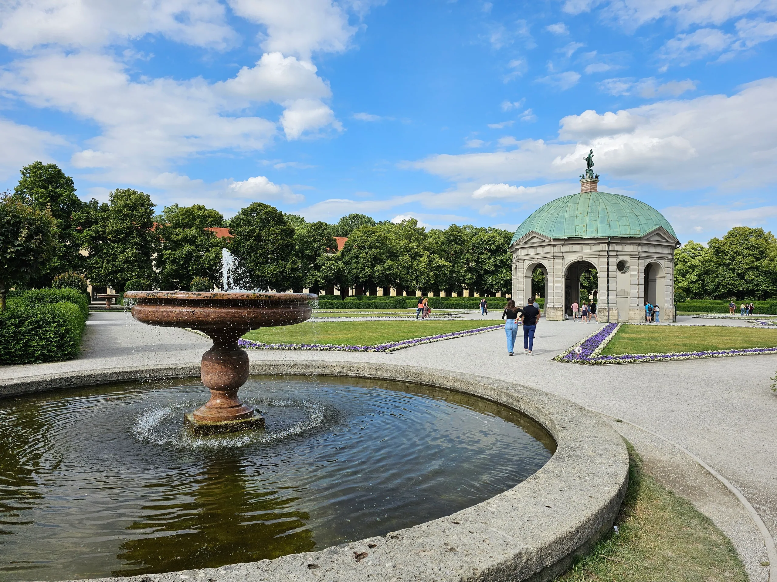 A stone fountain playing in a round basin in the Hofgarten, the domed Diana temple and clipped hedges beyond.