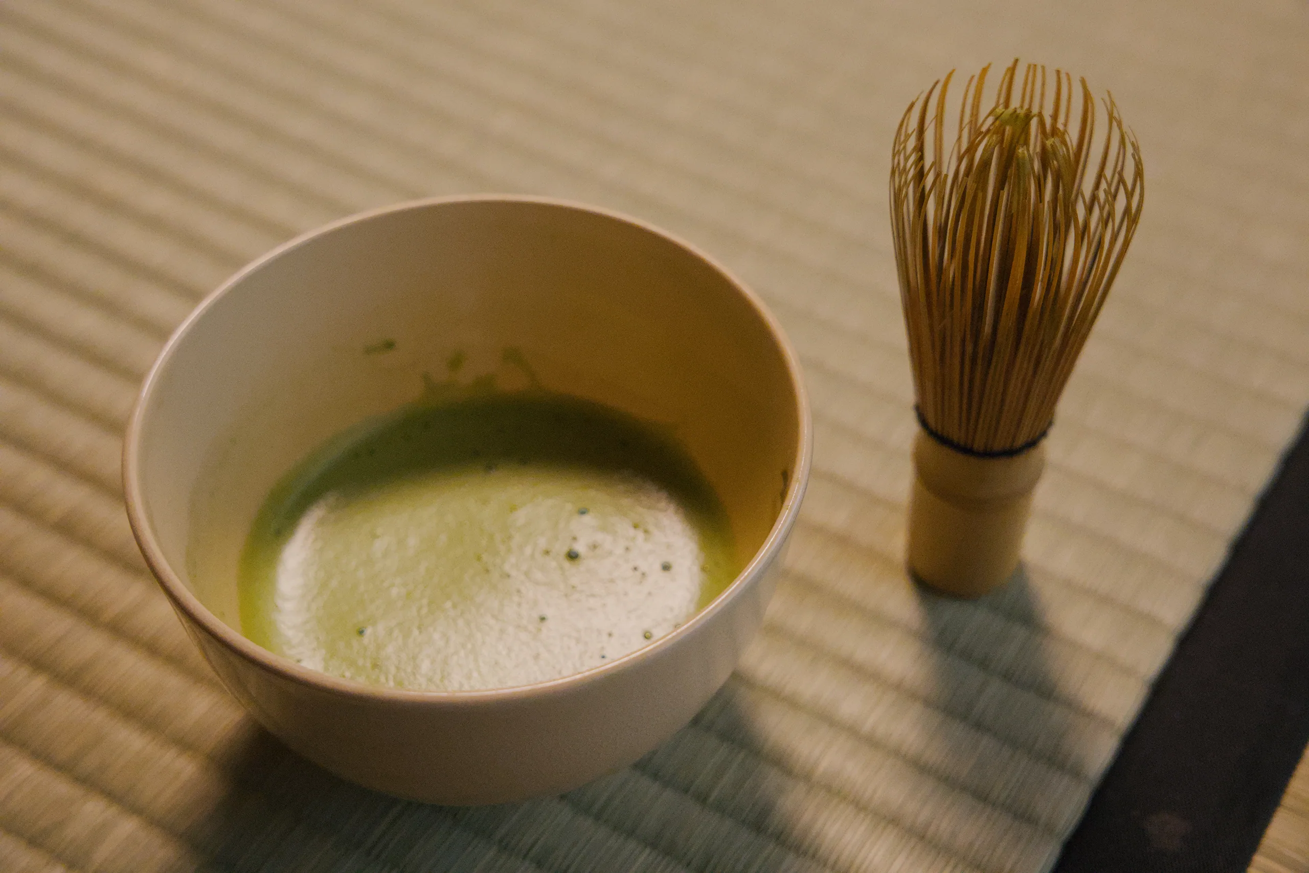 A white bowl of matcha tea with a bamboo whisk beside it.