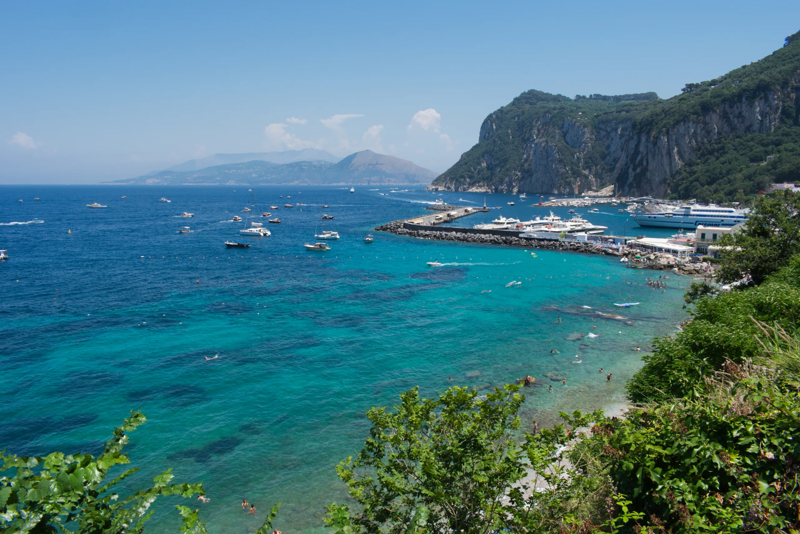 The harbour at Capri from above, boats moored along the breakwater and clear turquoise water below steep green cliffs.