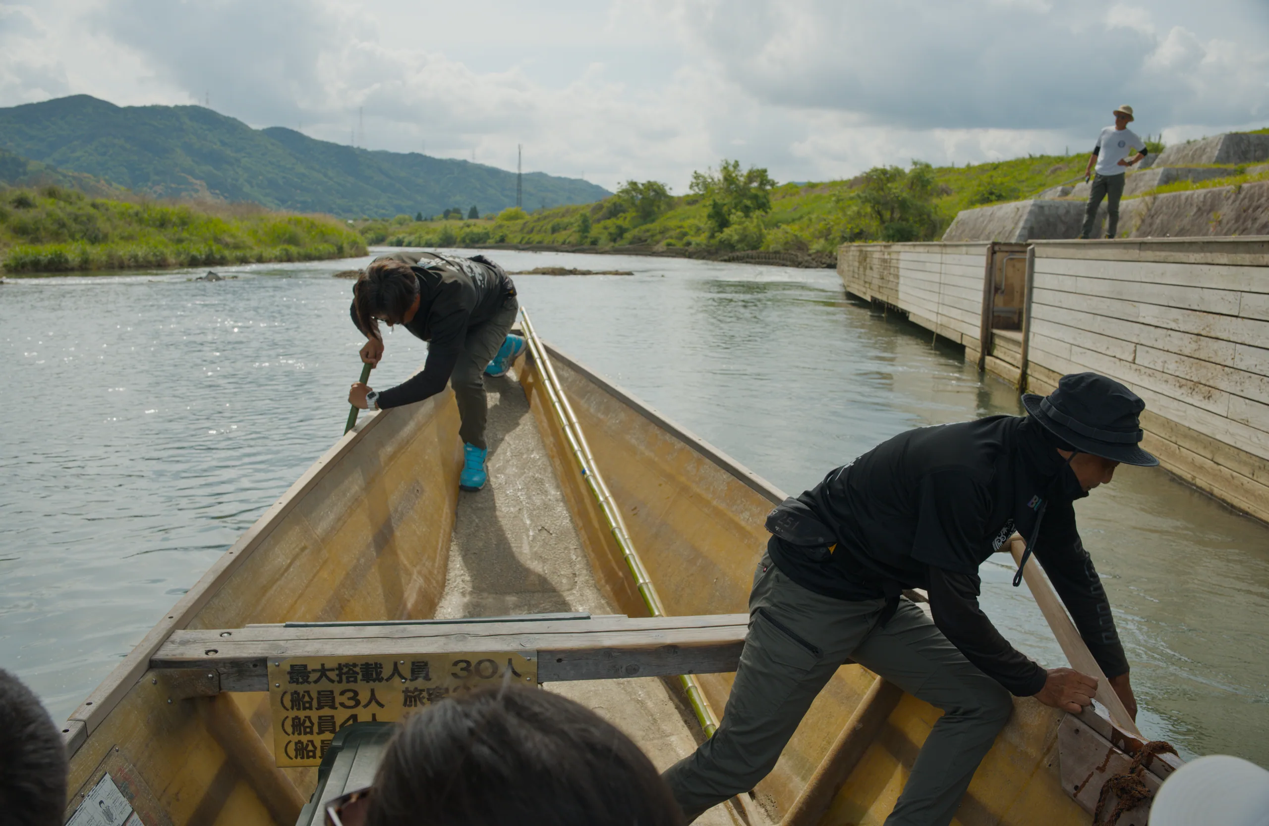 Boatmen propel a traditional wooden boat upriver with mountains visible in the distance.