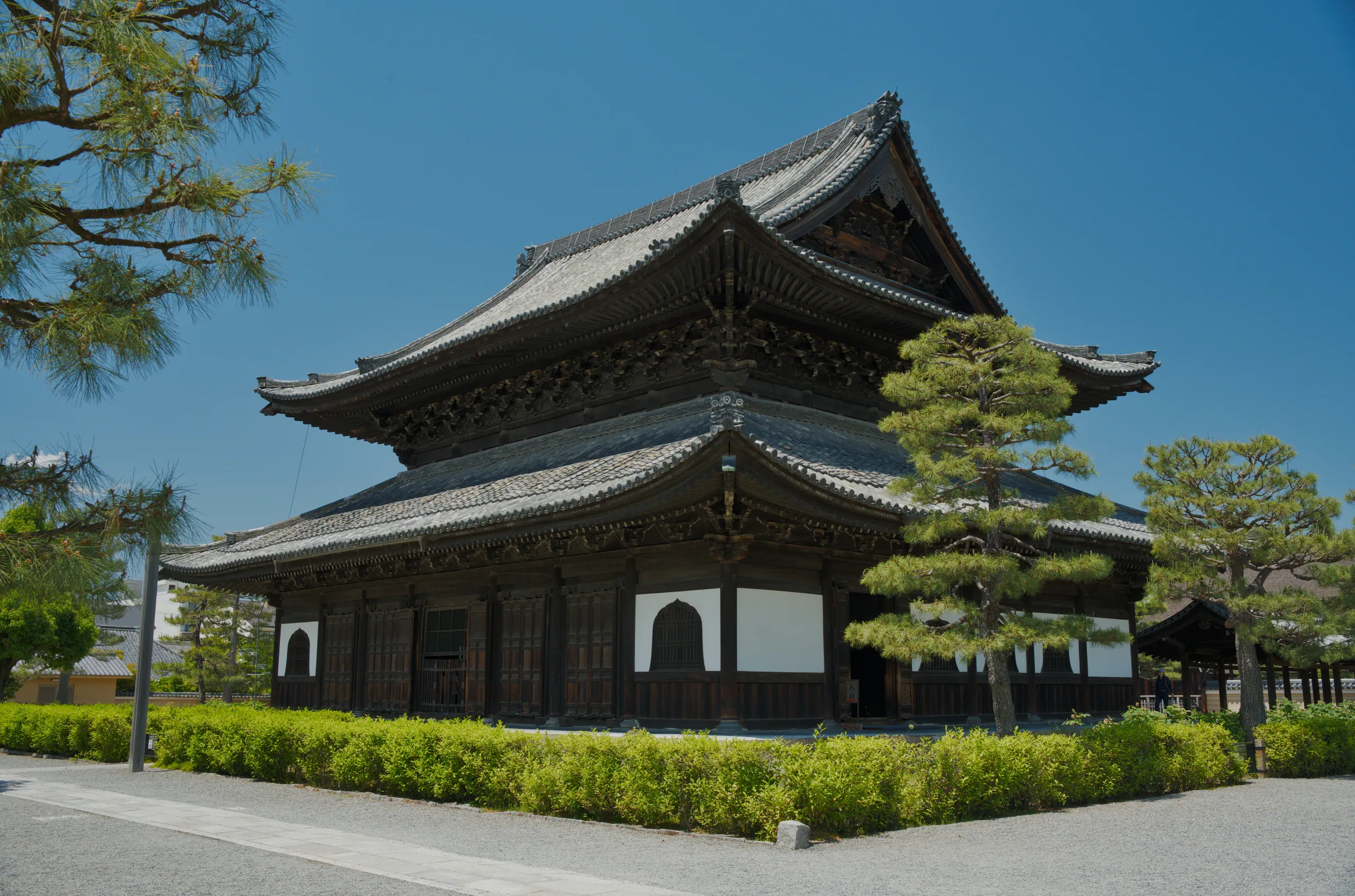 A traditional East Asian temple pavilion with upturned eaves stands in a manicured garden with specimen trees and hedges.