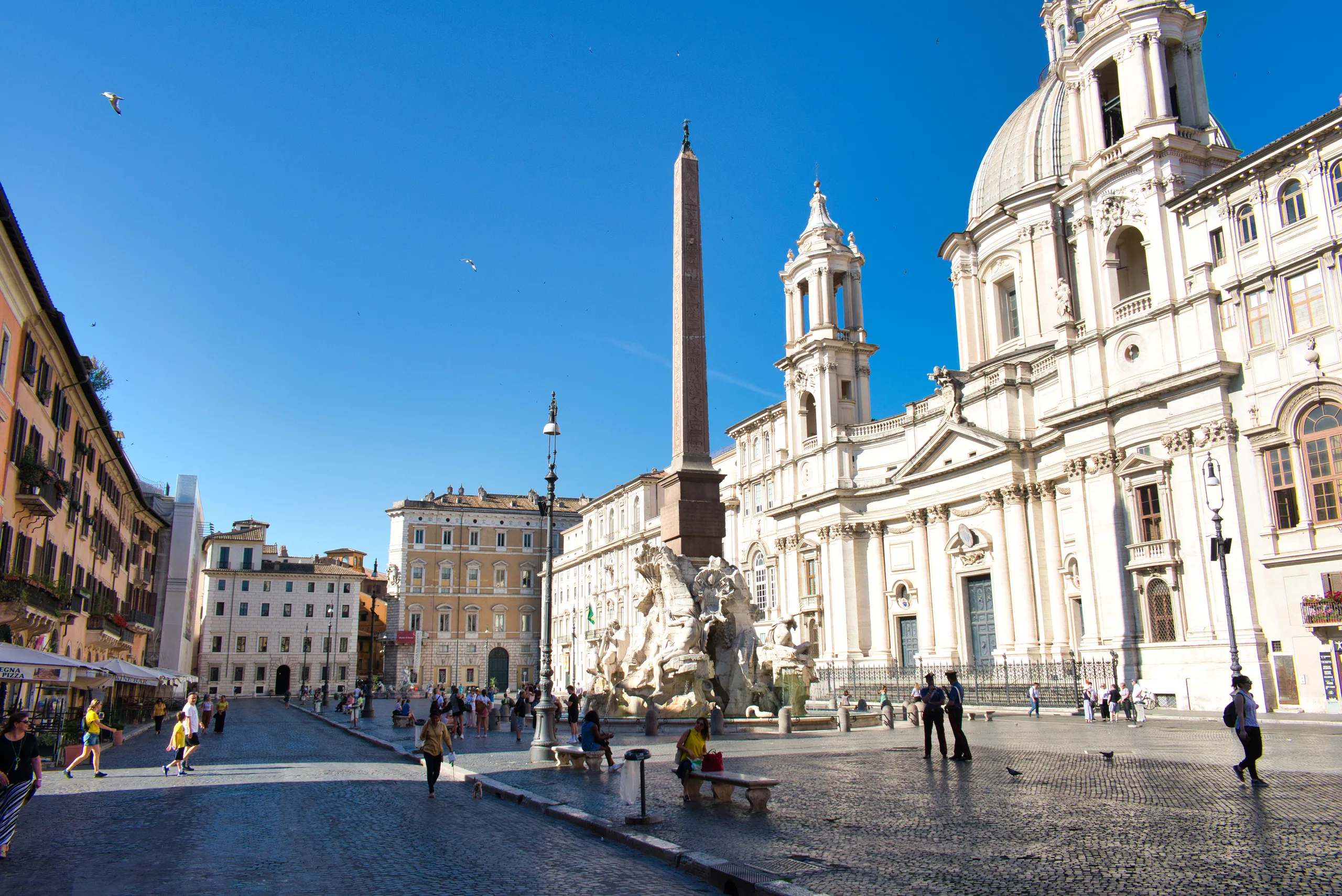 Piazza Navona in Rome, the obelisk and Sant'Agnese in Agone above the square, people crossing the paving in bright sun.