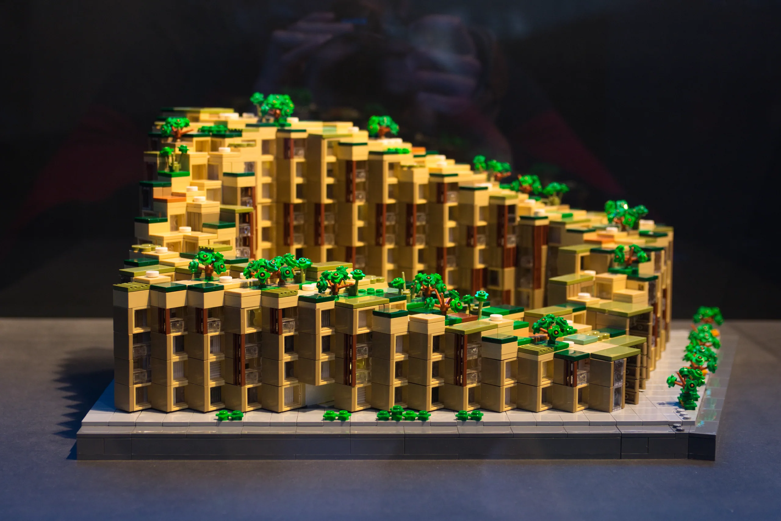A detailed architectural model built from yellow LEGO bricks, stepped like a terraced housing block and planted with small green trees.