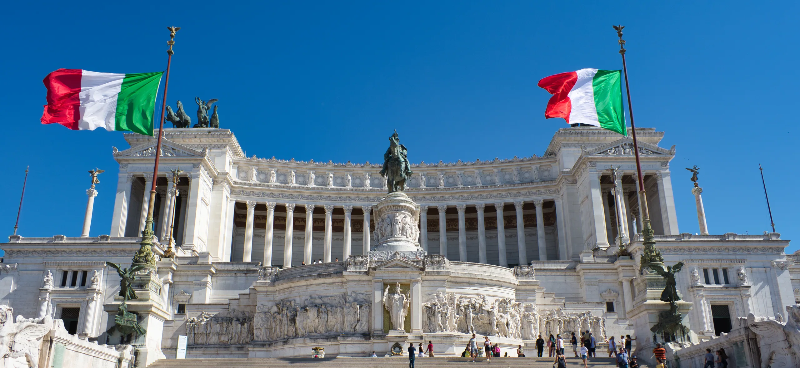 The white marble Vittoriano monument with Italian flags flying at either end against a deep blue sky.