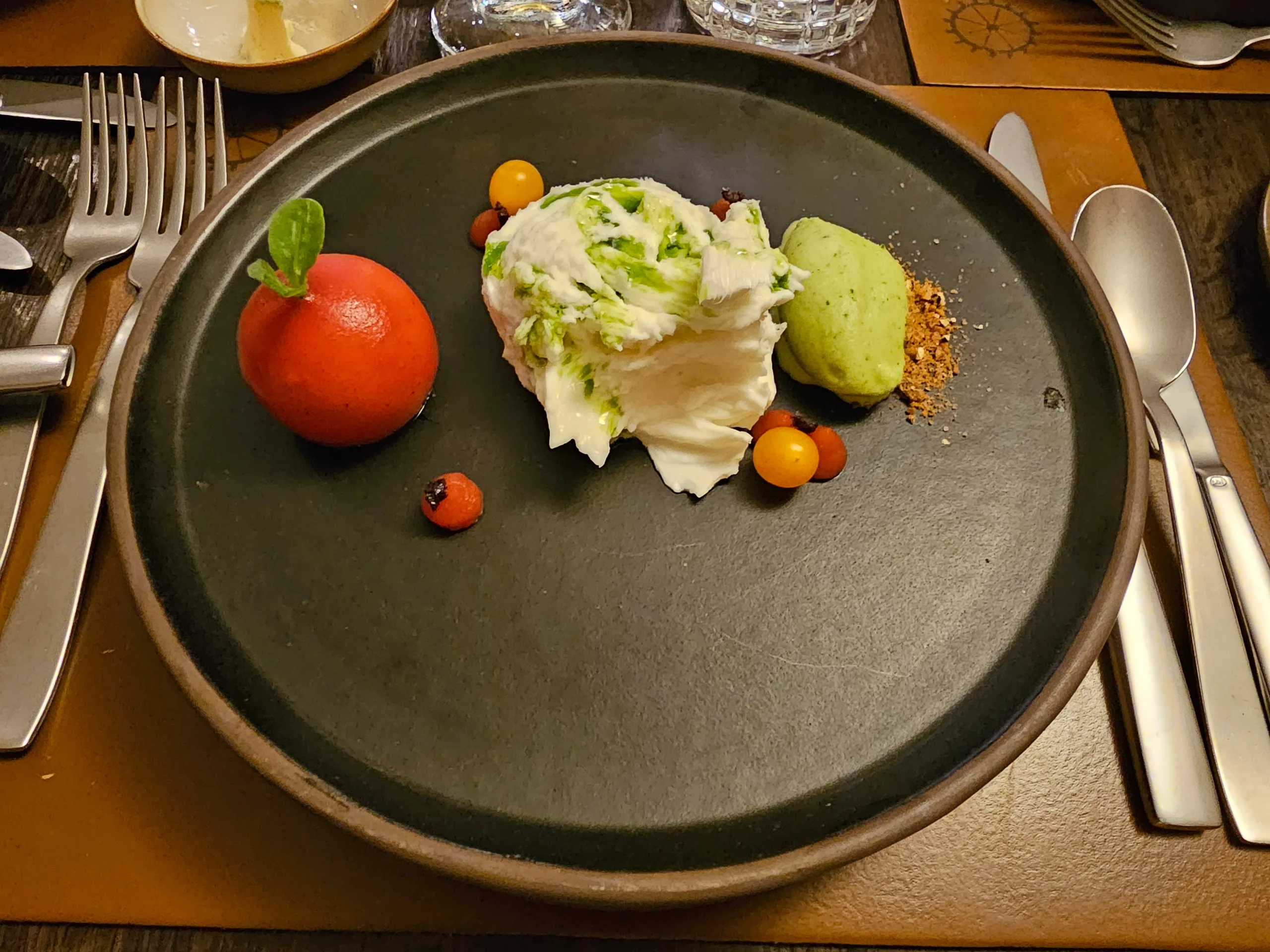 A plated course of whipped cheese with herbs, a bright orange tomato and small yellow tomatoes on a dark plate.