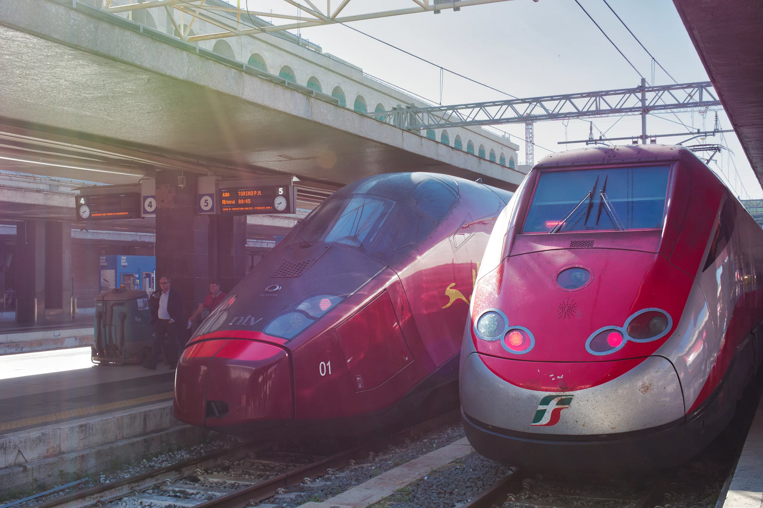 Two red Frecciarossa high-speed trains standing nose to nose at a station platform under a glazed roof.
