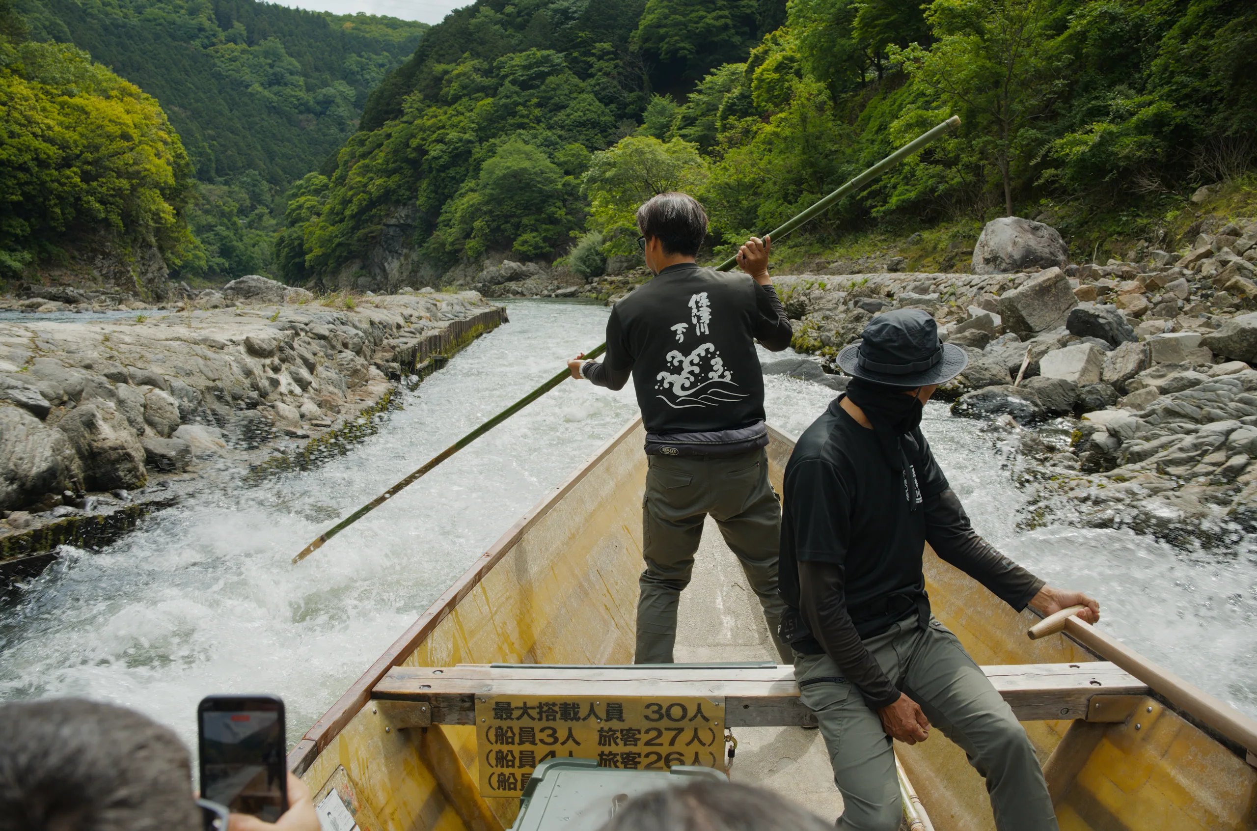 Two people stand in a yellow boat on a rocky river beneath forested mountains.