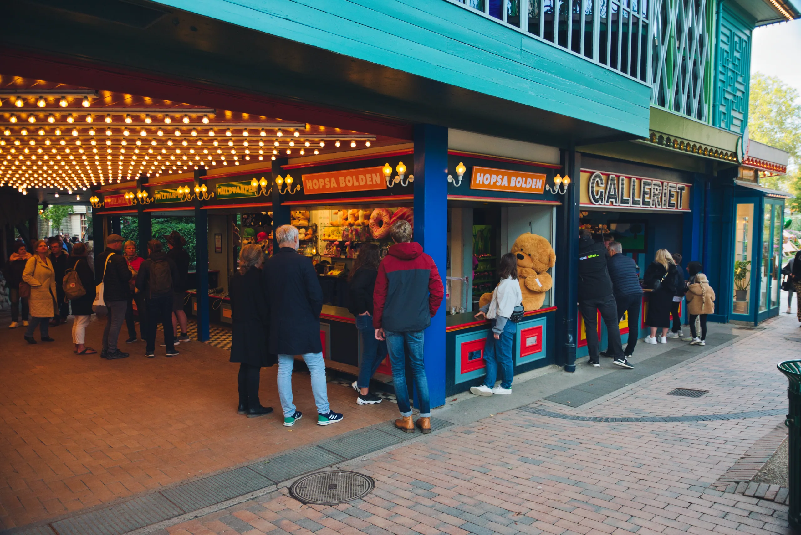 A row of brightly lit game stalls at Tivoli, people queueing along a curved arcade under strings of bulbs.