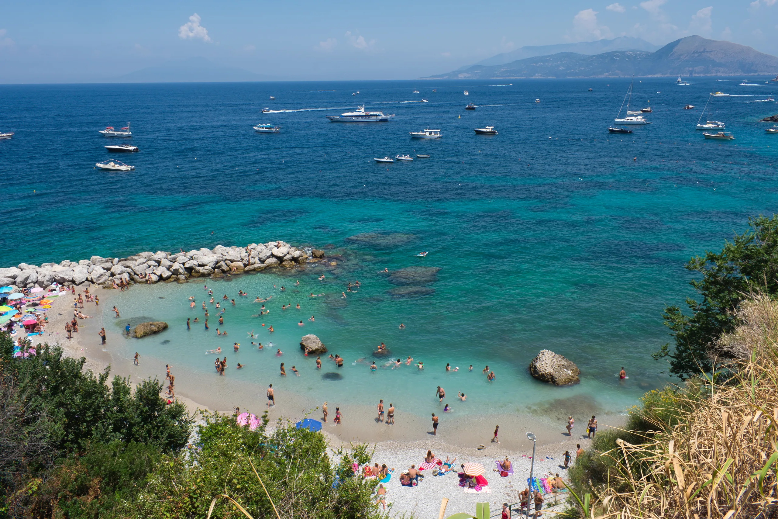 A crowded beach in a small cove, sunbathers and umbrellas along the sand and boats anchored in the turquoise water beyond.