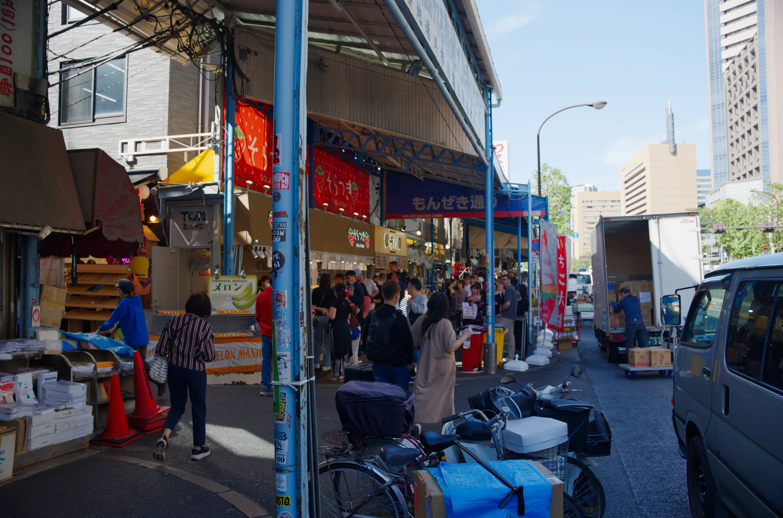 Shoppers walk past storefronts on a crowded street with blue structural columns and overhead signage.