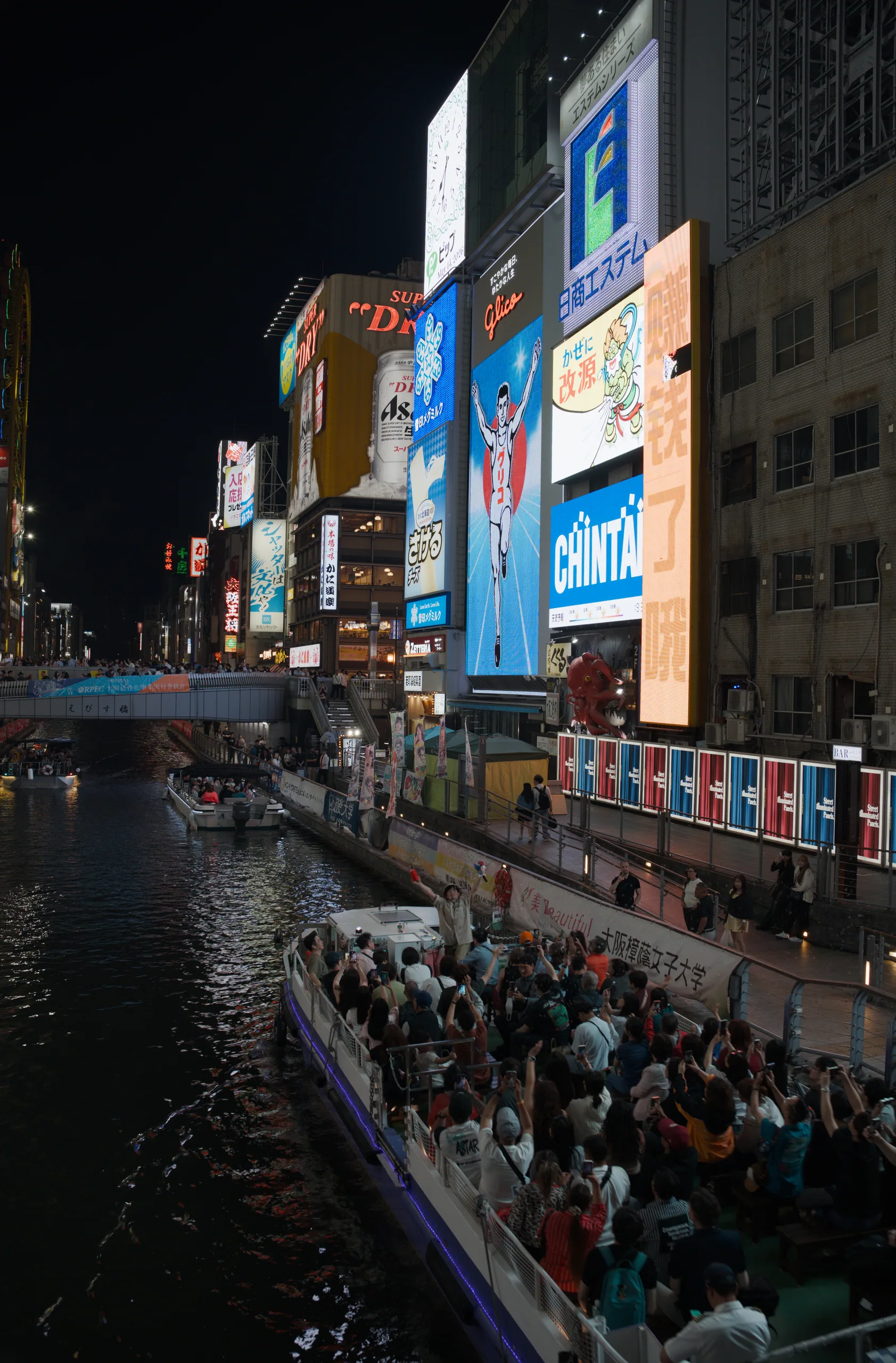 Neon signs and buildings reflected in a canal at night with crowds and bright storefronts.