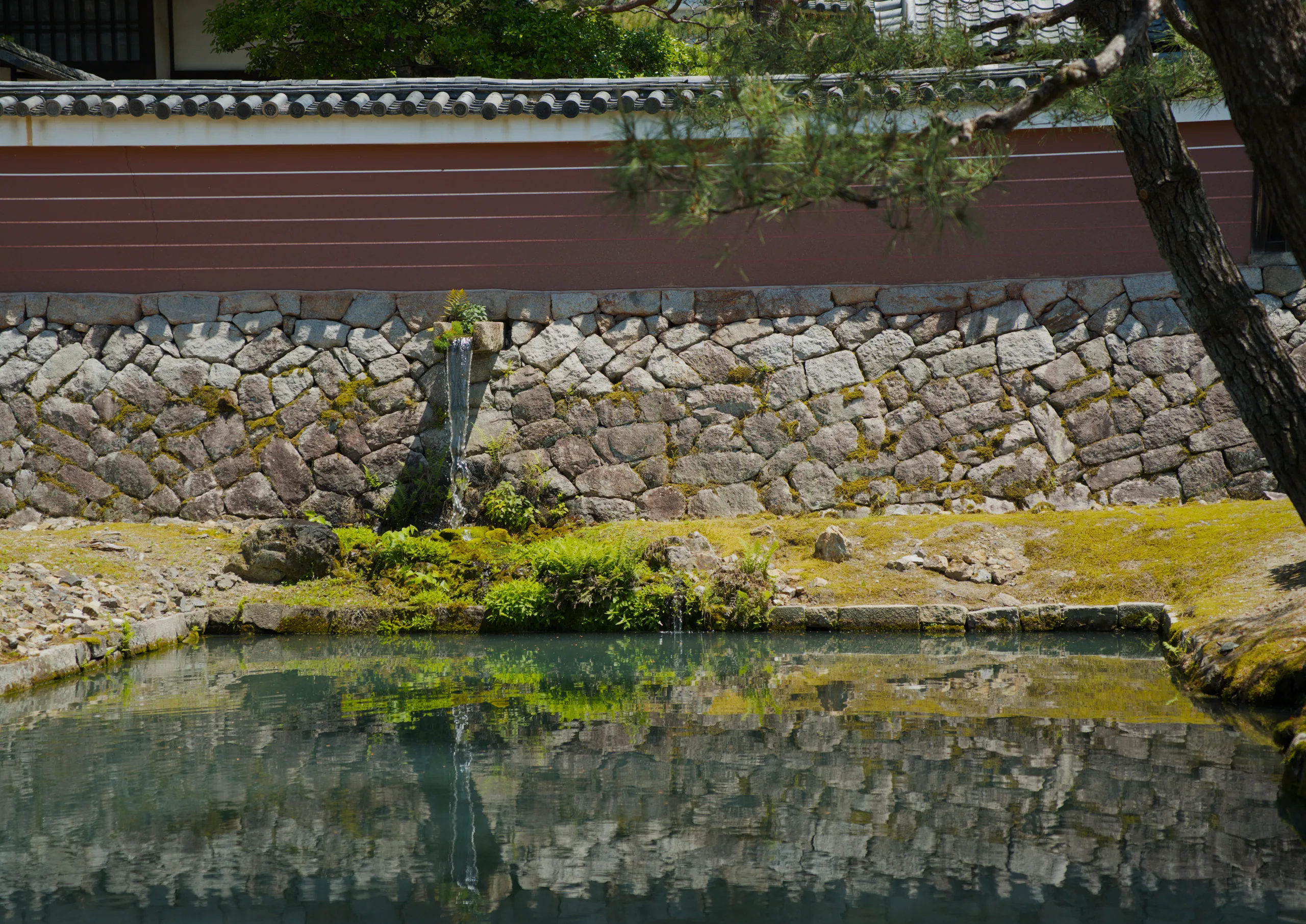 A massive stacked-stone fortification wall is reflected in still water surrounded by moss and manicured vegetation.