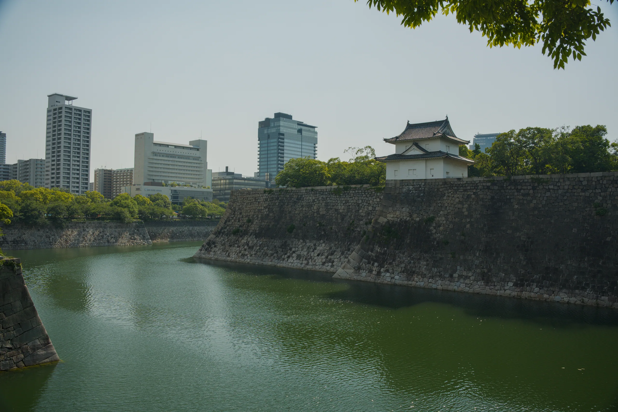 Japanese castle towers with curved rooflines stand above a wide moat bordered by steep stone walls.