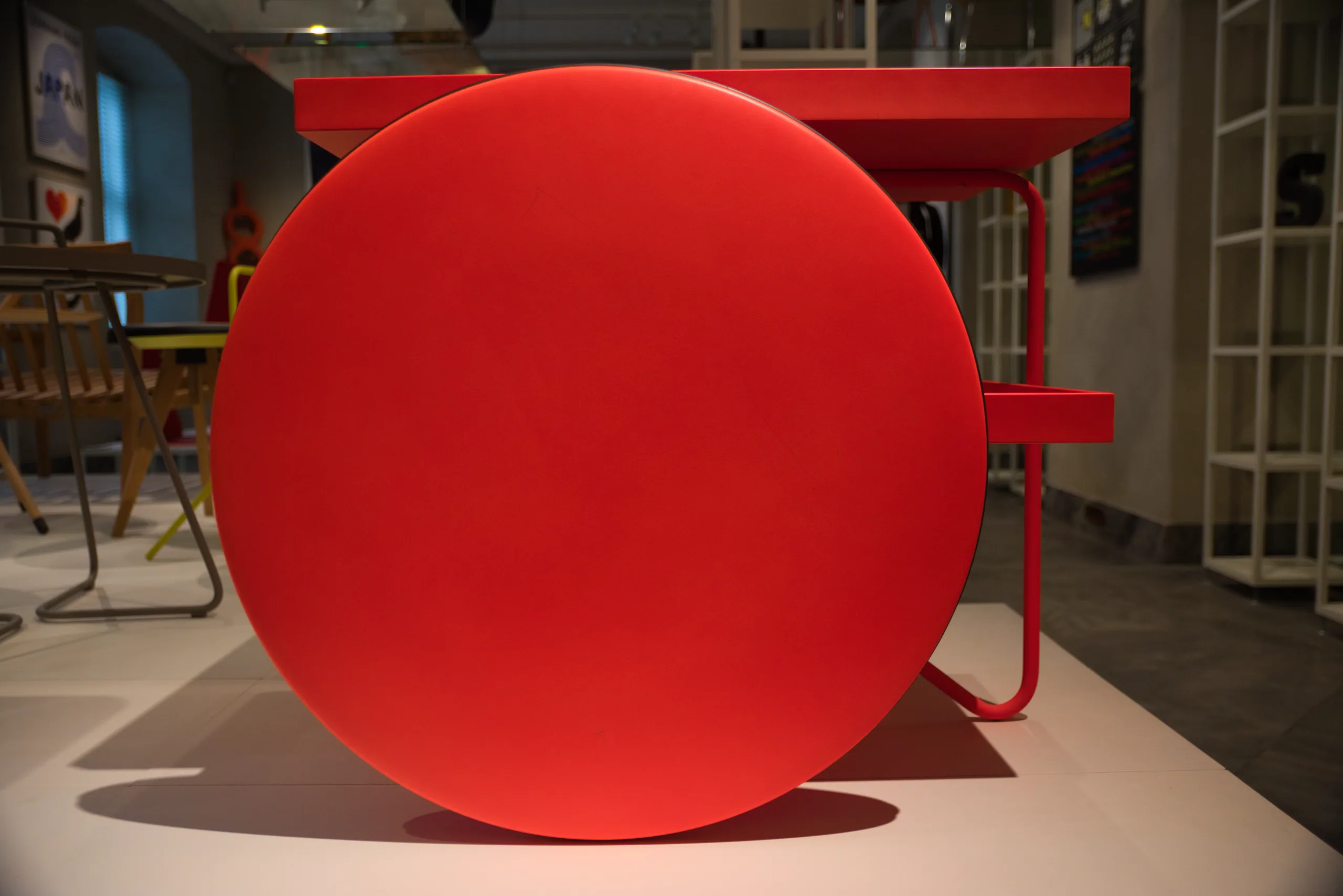 A bright red disc-shaped side table on a plinth in a design gallery, chairs and displays behind it.