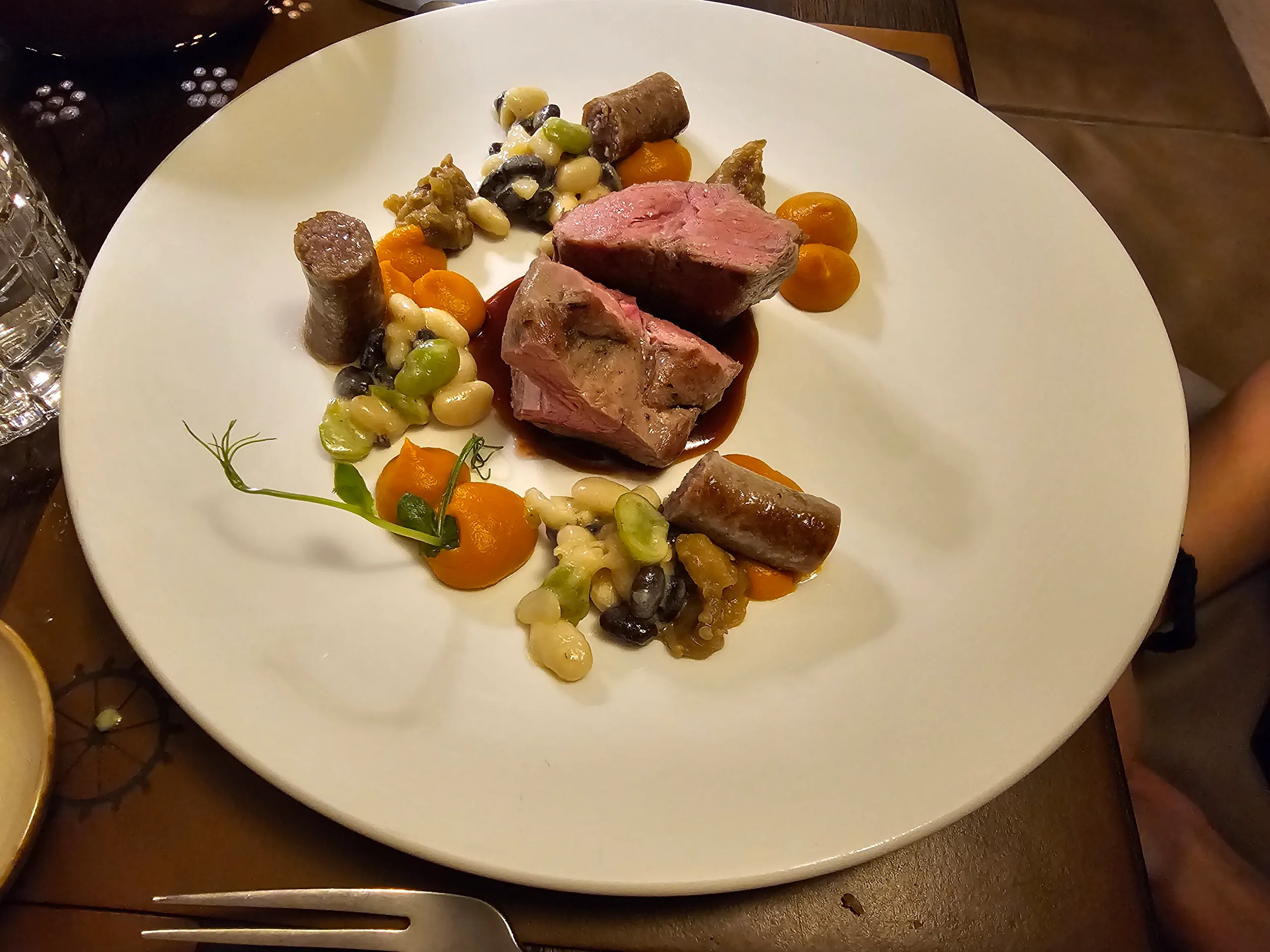 Slices of pink lamb with sausage, beans, carrot puree and herbs arranged around a white plate.