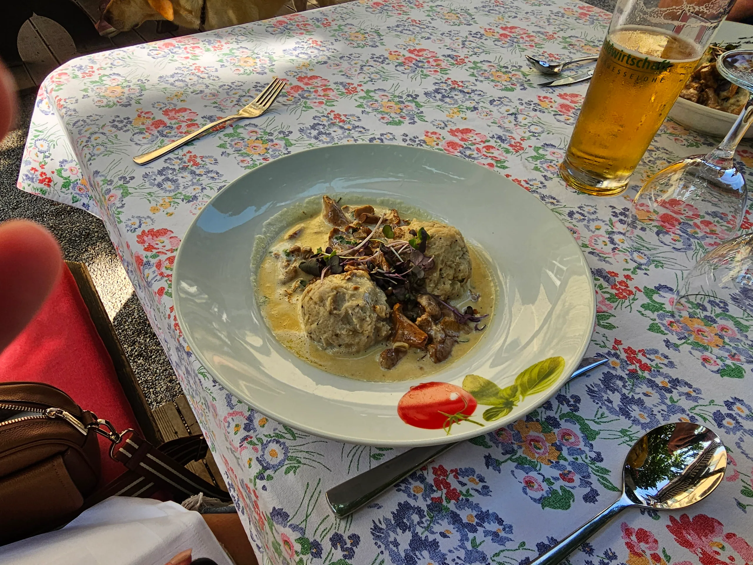 Bread dumplings in a creamy mushroom sauce on a floral tablecloth, a glass of beer alongside.