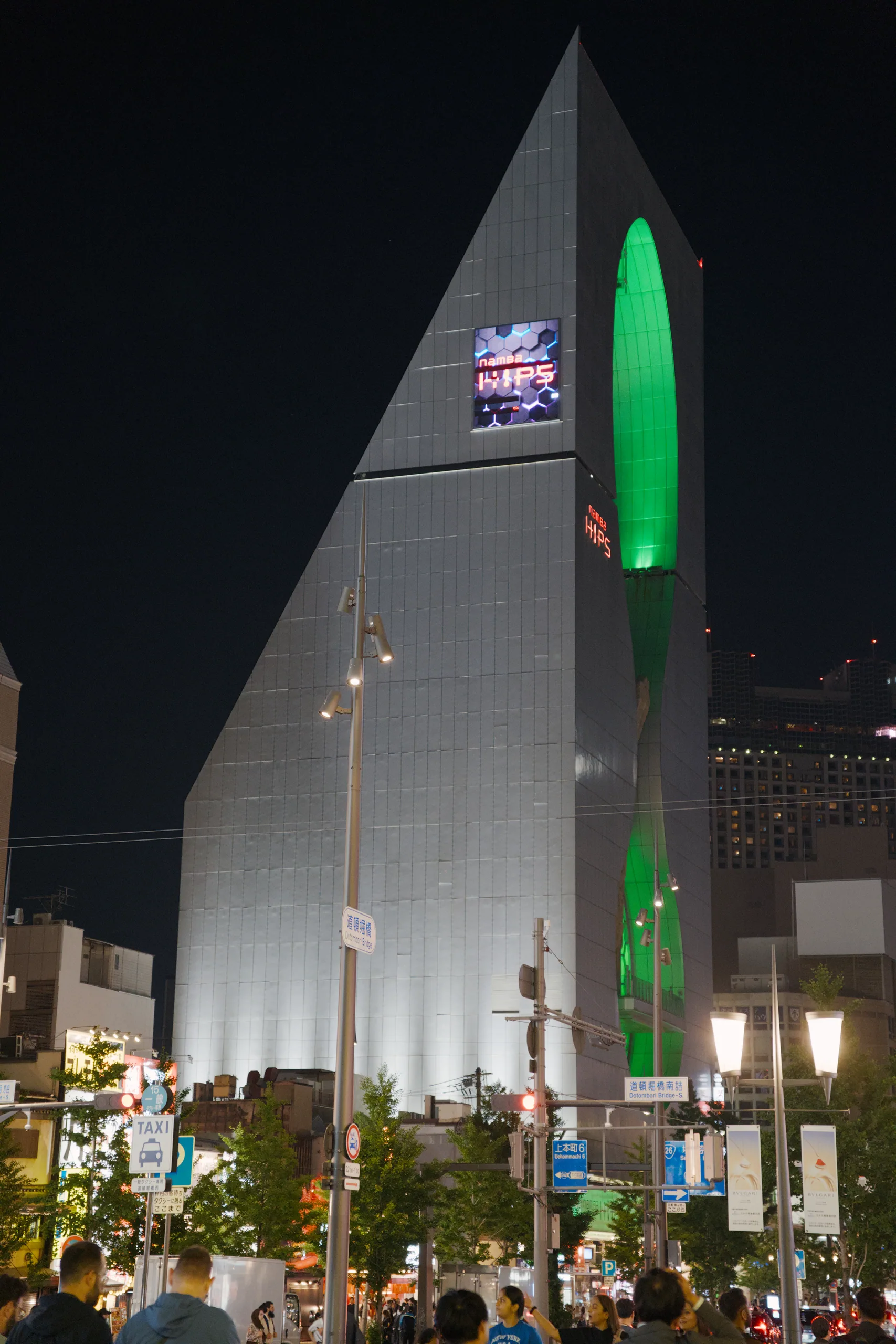 A conical white monument with a bright green neon element illuminated against the night sky, surrounded by crowds.