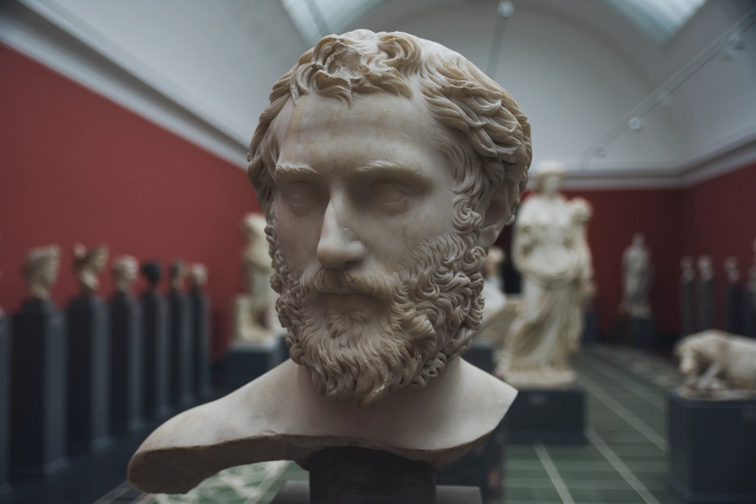 A marble portrait bust of a bearded man in a red-walled gallery, rows of classical statues receding behind it.