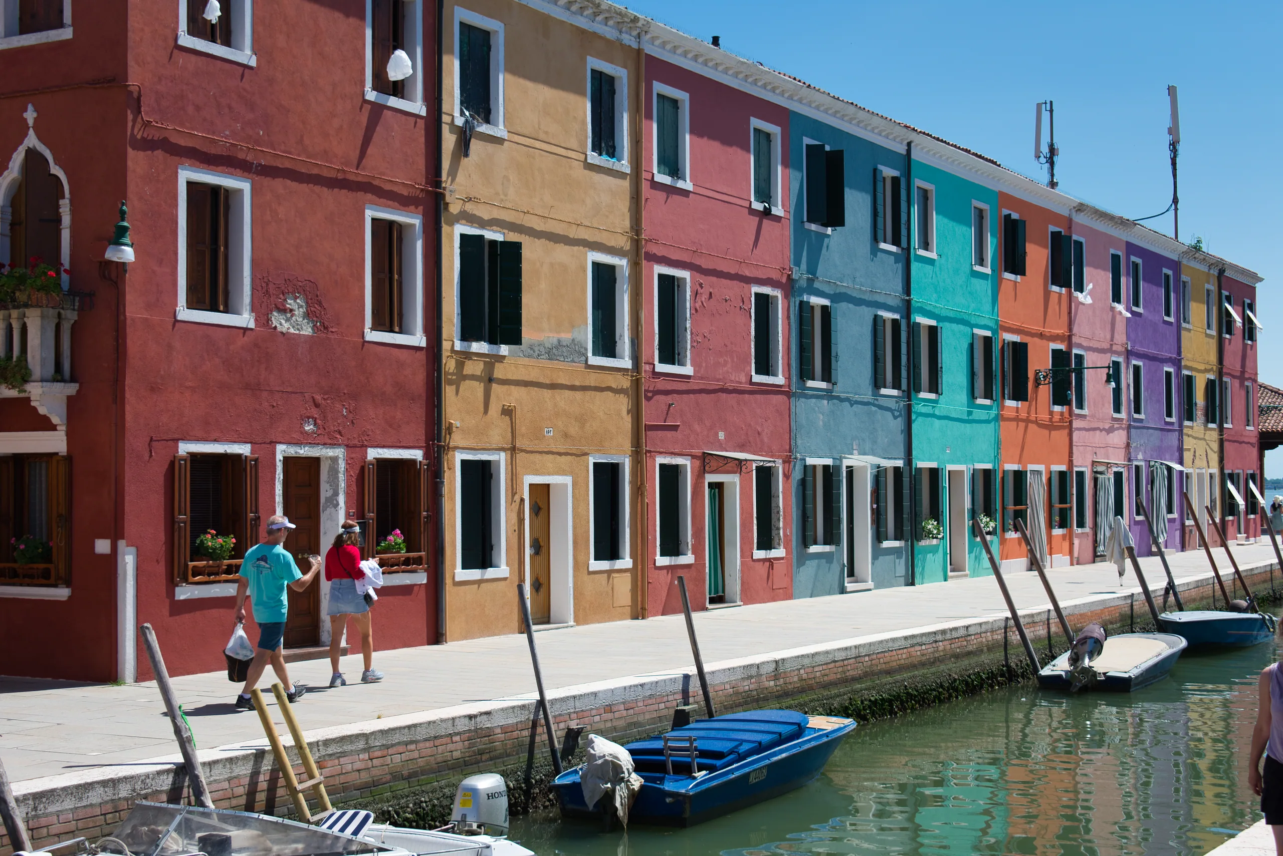 A row of Burano houses in ochre, red, blue and green along a canal, small boats moored in front and people walking the quay.