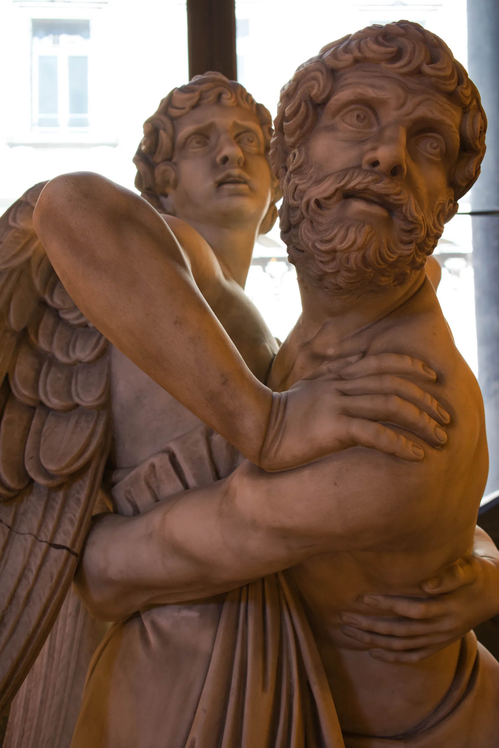 A terracotta sculpture of a winged angel embracing a bearded man, lit warmly against a pale wall.