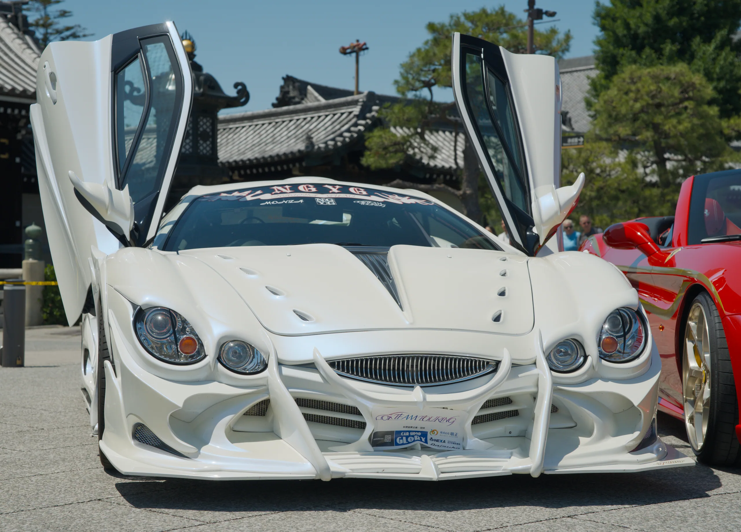 A white sports car displays with butterfly doors opened at a car show, with a red supercar visible beside it.
