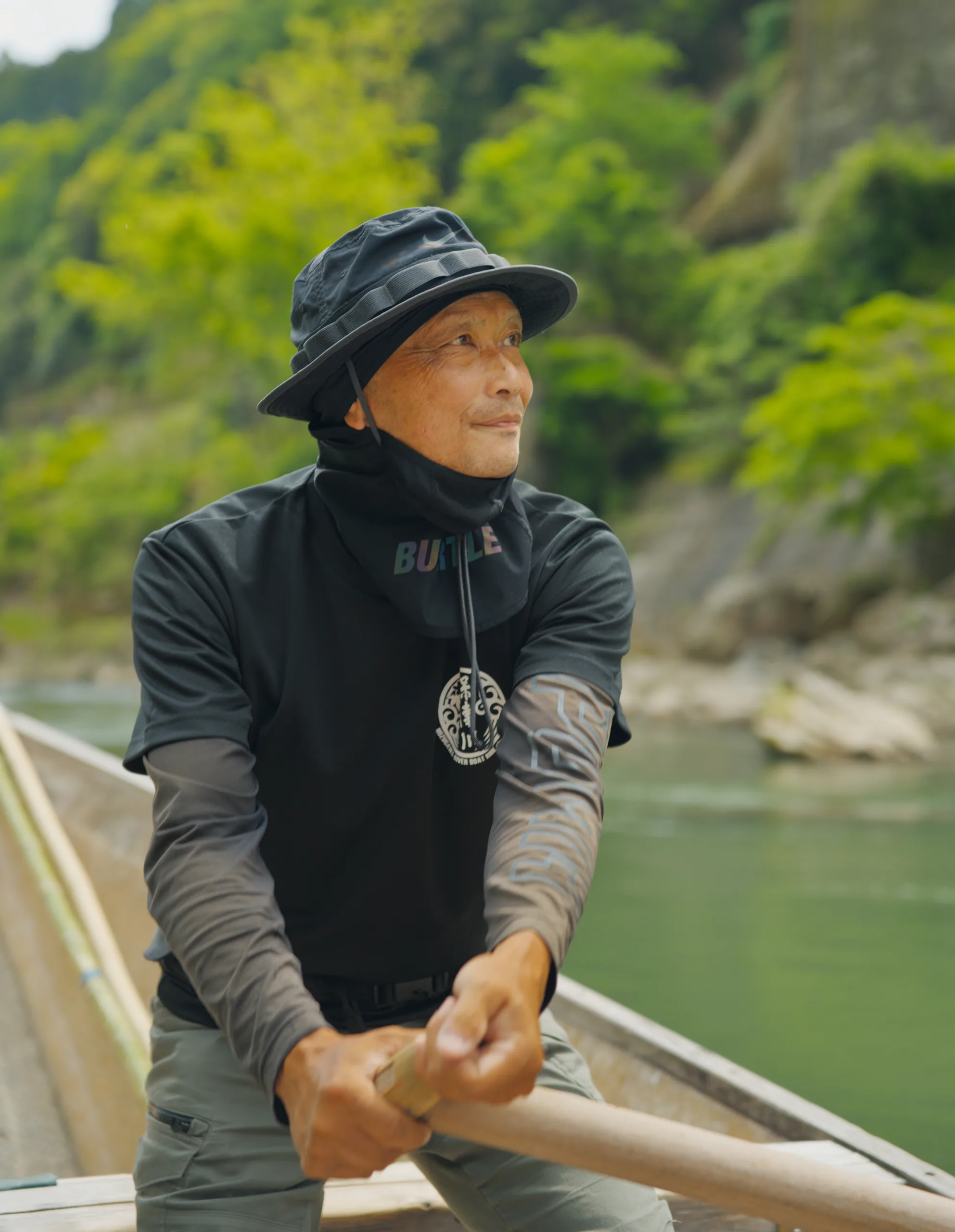 Person in a hat seated in a boat on calm river water with forested hills in the distance.