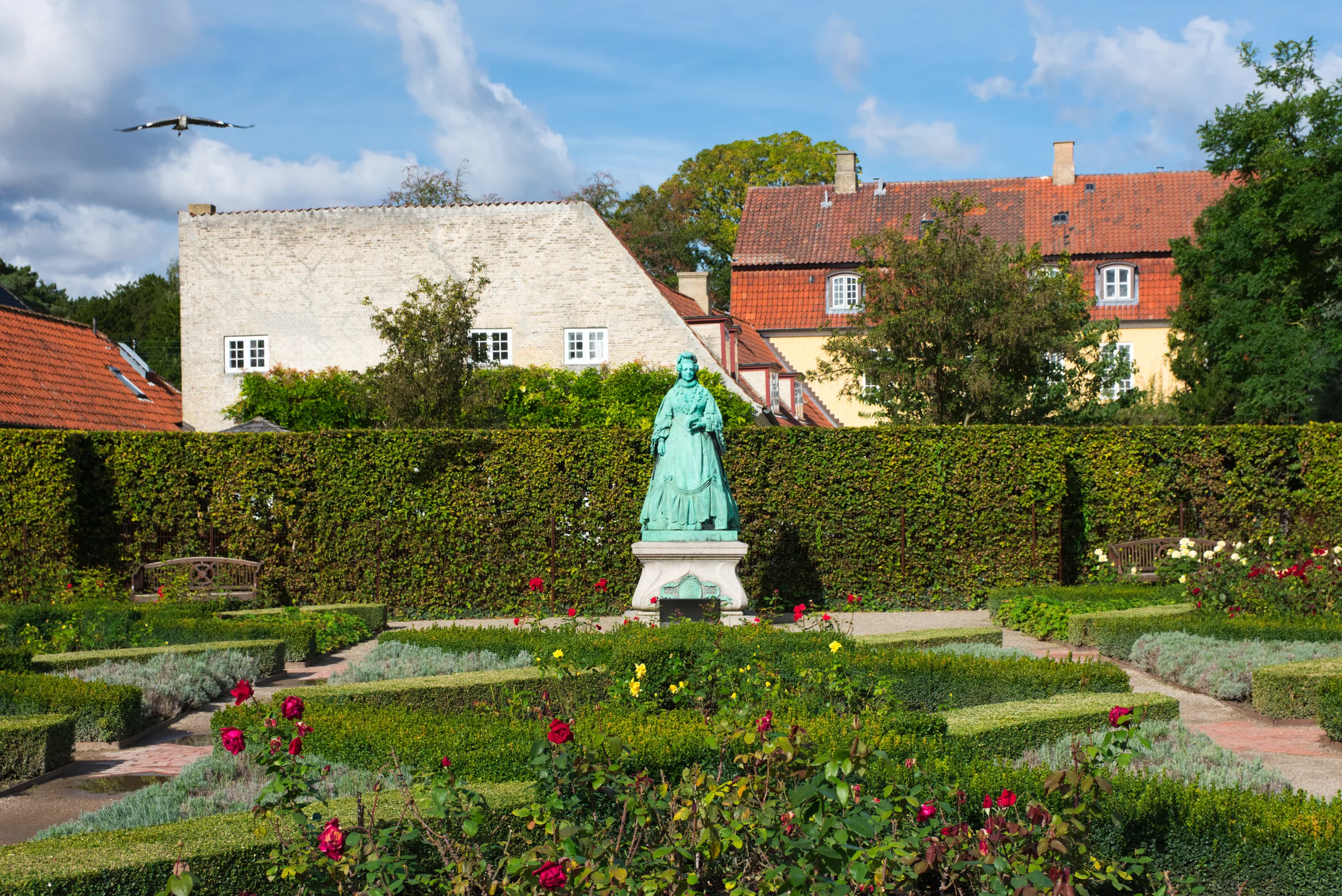 A formal rose garden of low clipped hedges around a weathered green bronze statue, tiled roofs of surrounding buildings behind.