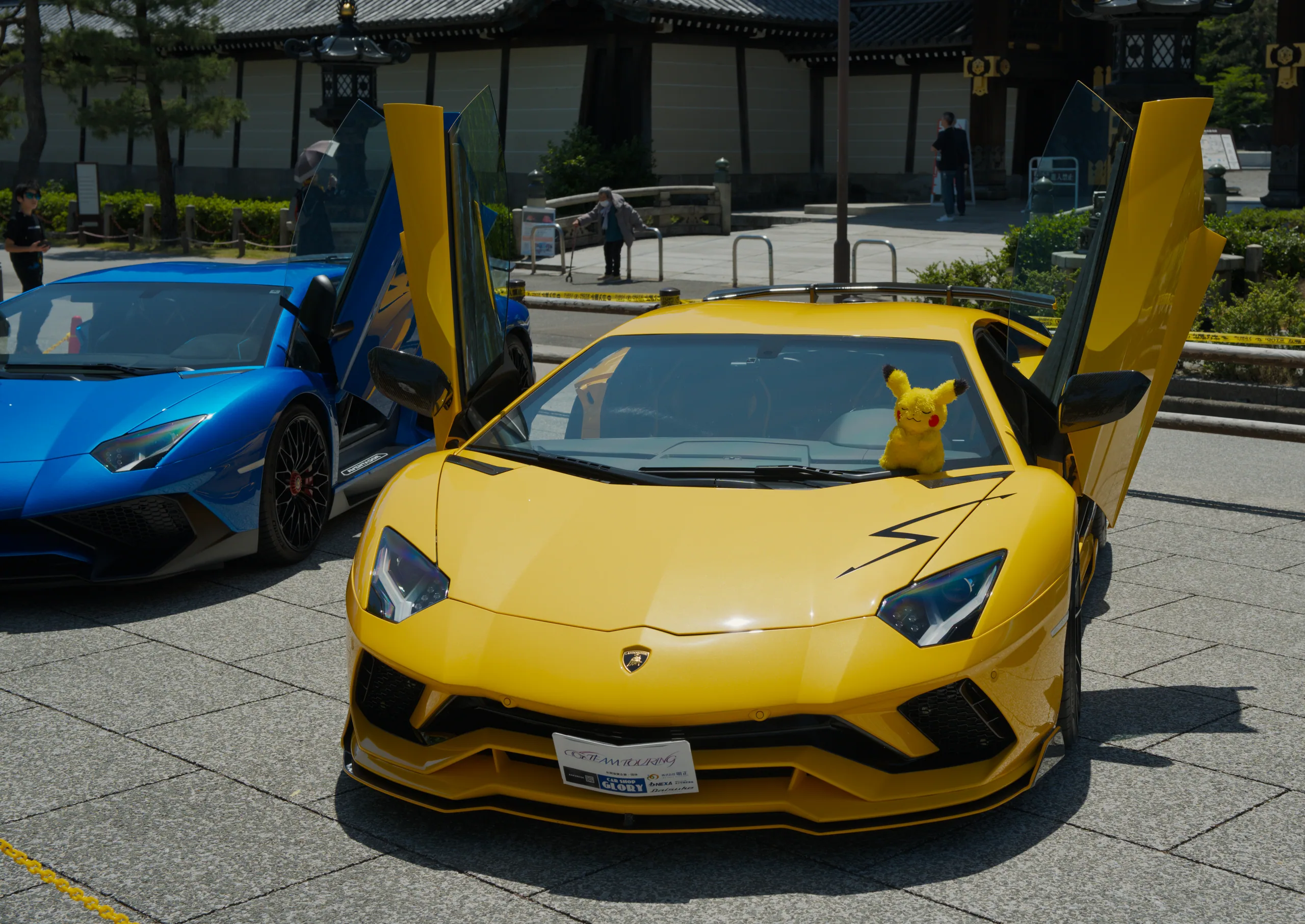 A bright yellow sports car with butterfly doors open, displayed at a car show with other vehicles around it.