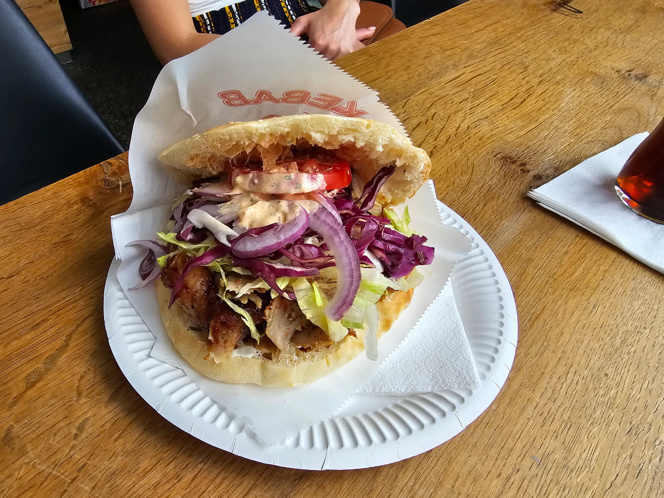 A doner kebab in flatbread with salad and red onion, on a paper plate on a wooden table.