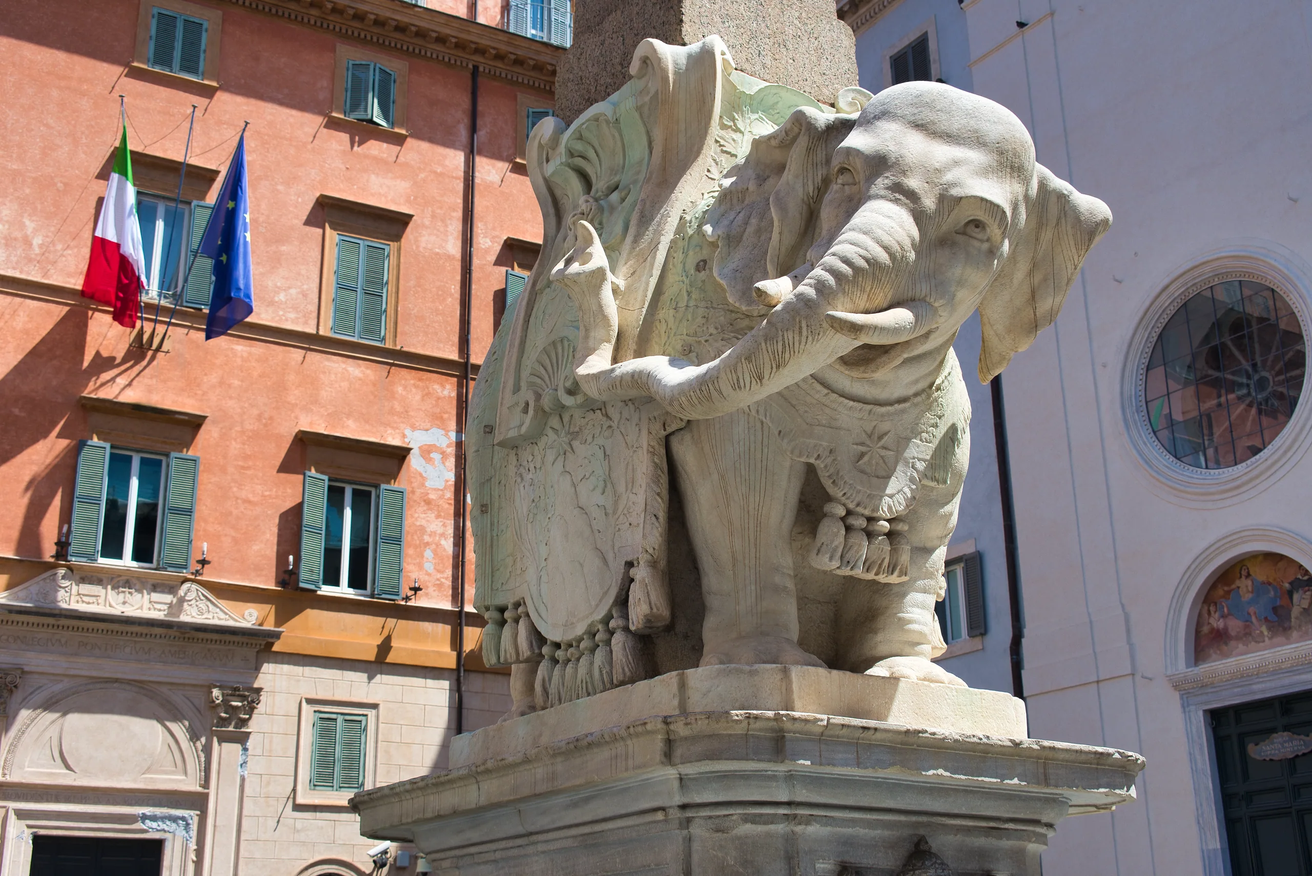 Bernini's marble elephant carrying an obelisk on its back, in front of an ochre facade hung with Italian and EU flags.