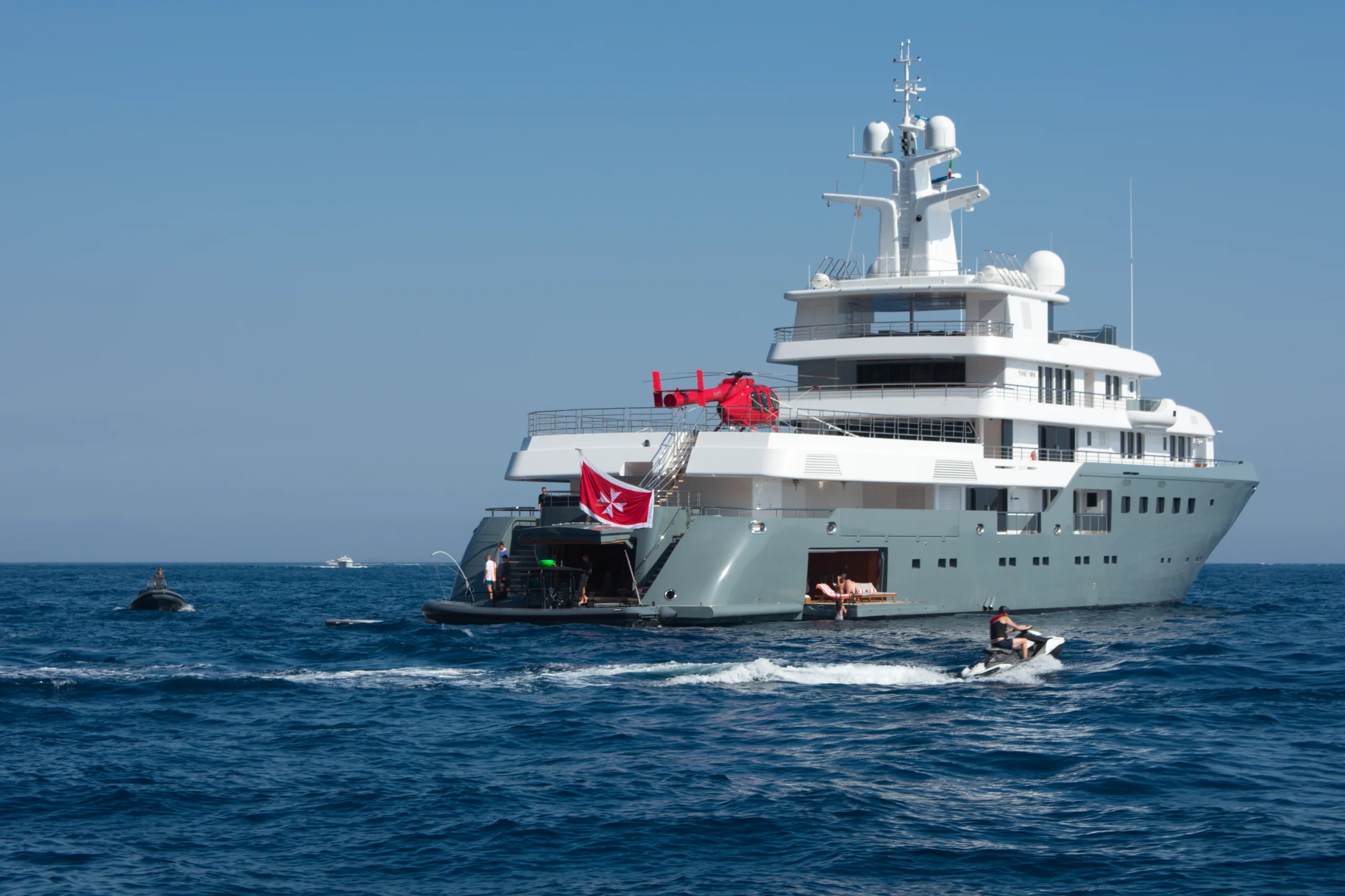 A superyacht at anchor on flat blue water, a red helicopter on its upper deck and a jet ski alongside.