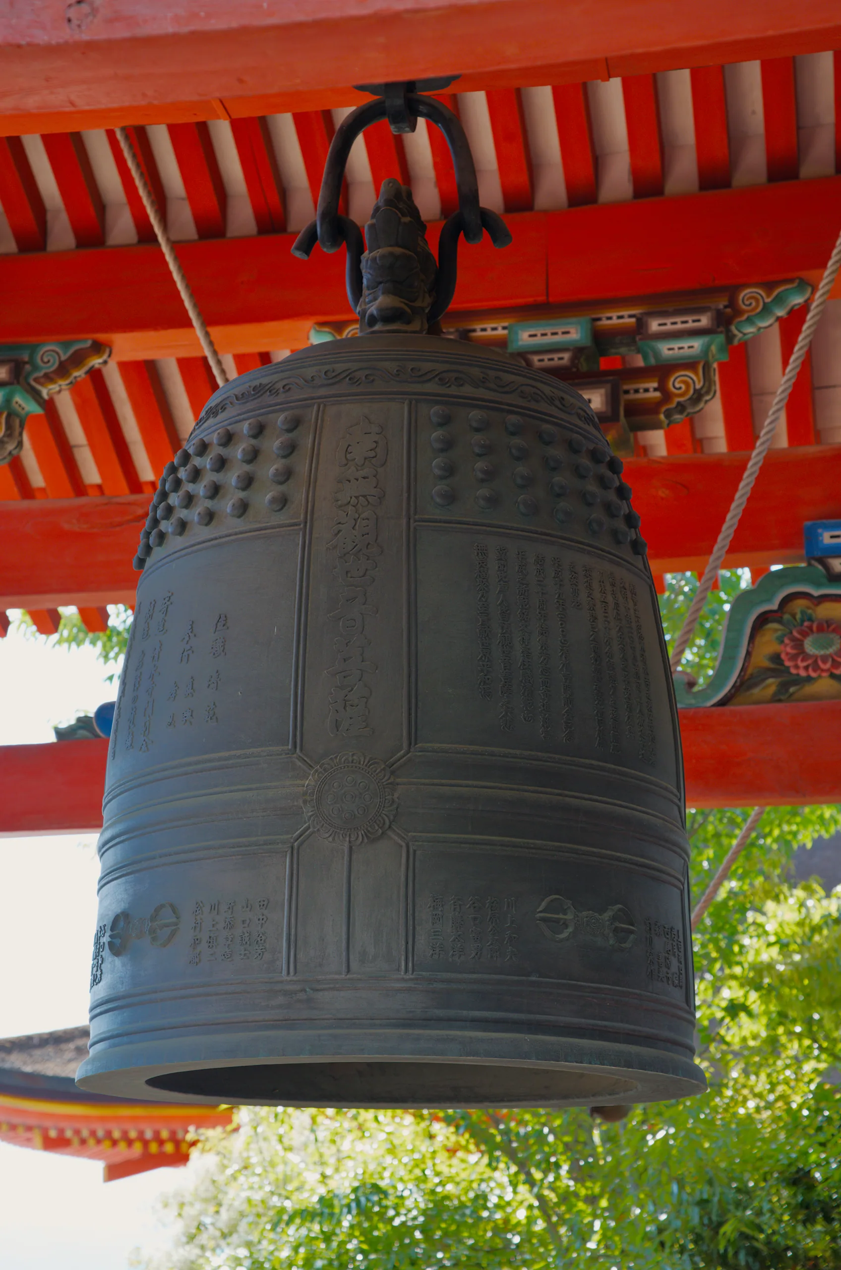A large bronze bell hangs from intricately carved wooden braces beneath bright red temple eaves.