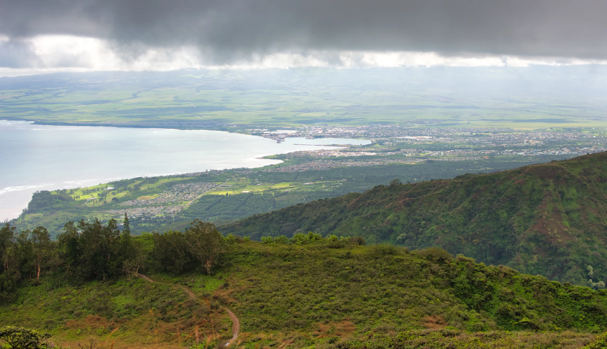 A high view over green ridges to a wide bay and coastal plain, a heavy grey cloud layer above.