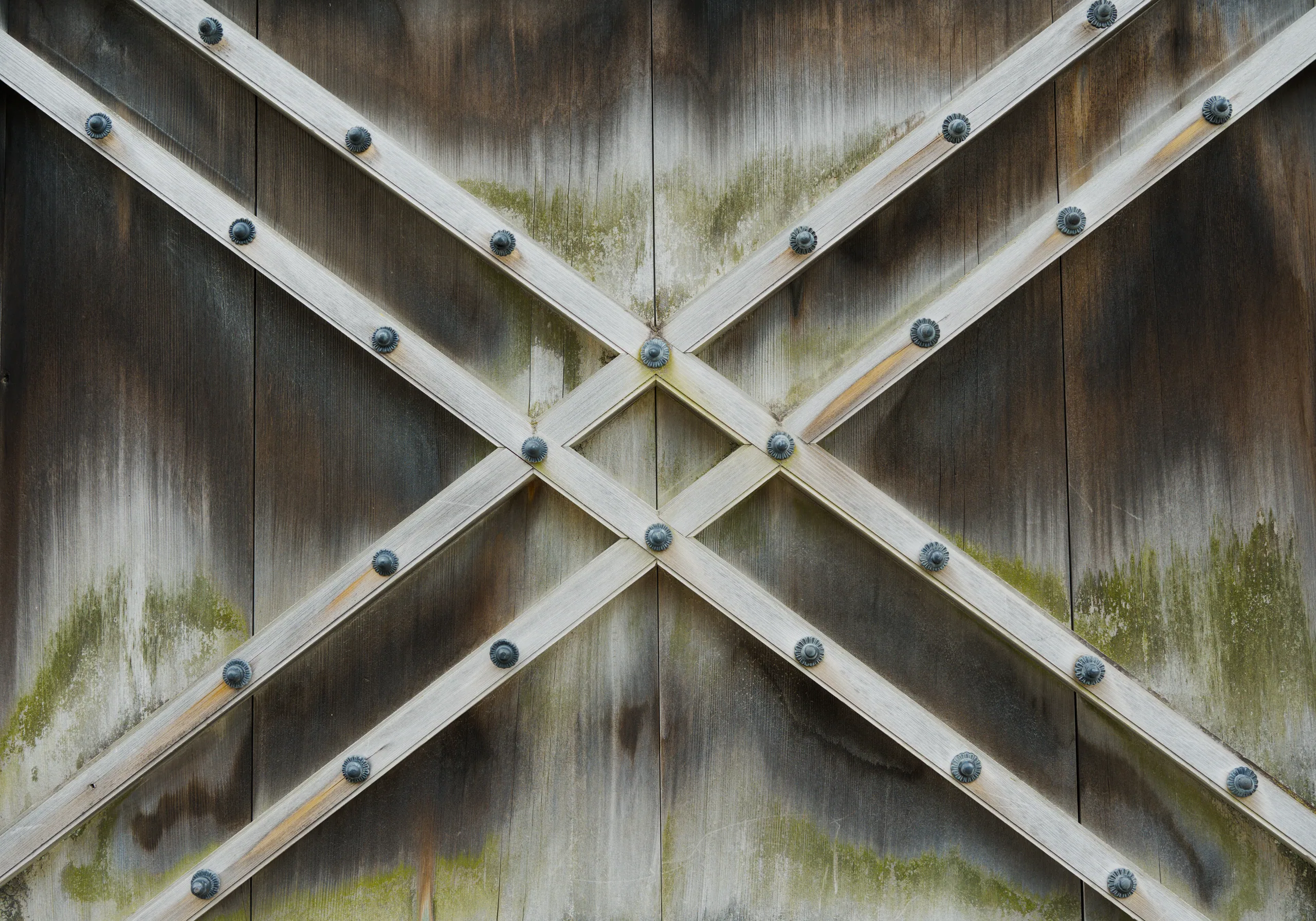Wooden architectural joinery with metal bolts in a cross pattern, surface weathered and moss-covered.