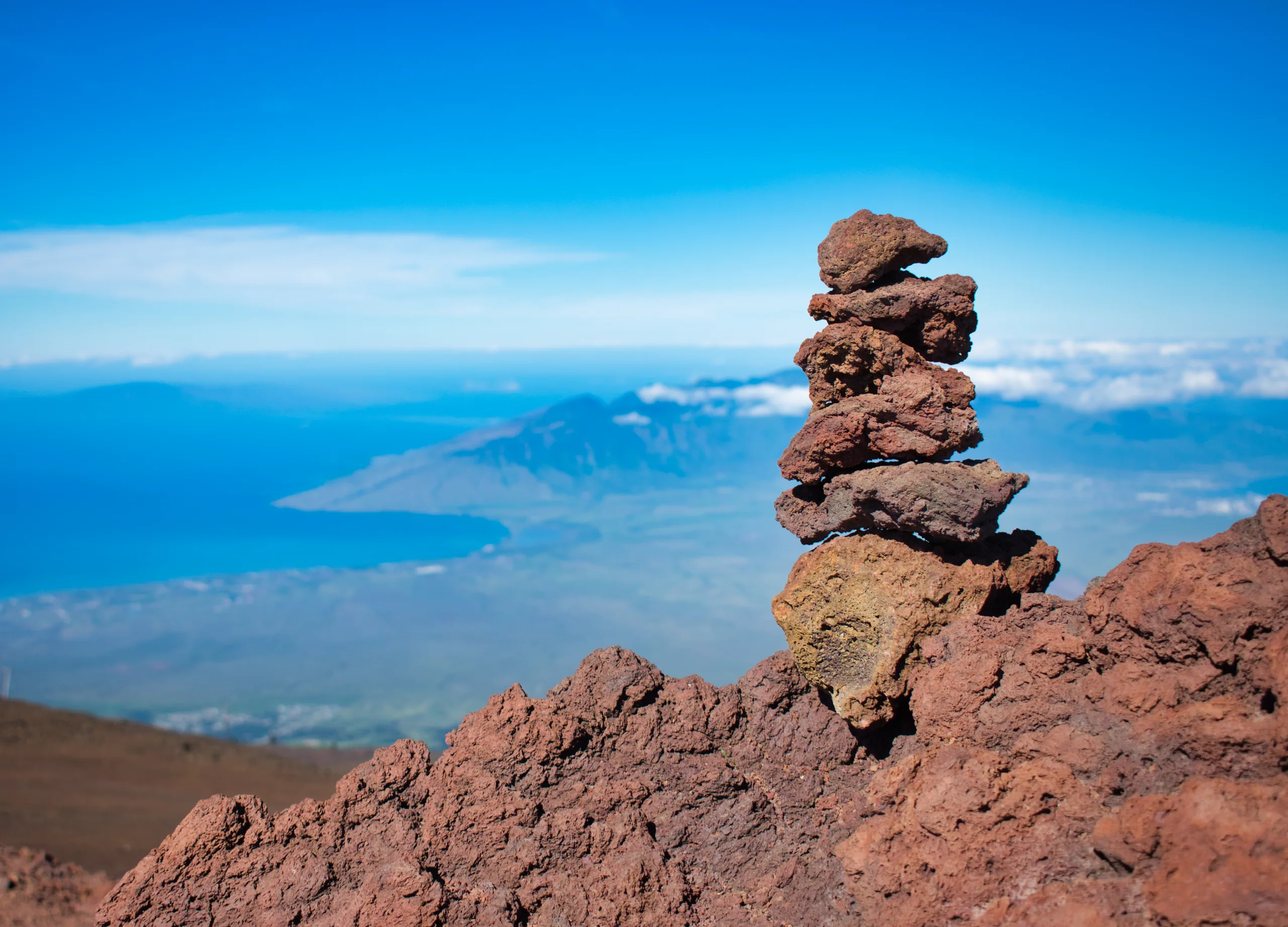 A stack of balanced red lava stones on a summit outcrop, with a cloud-fringed island far below.