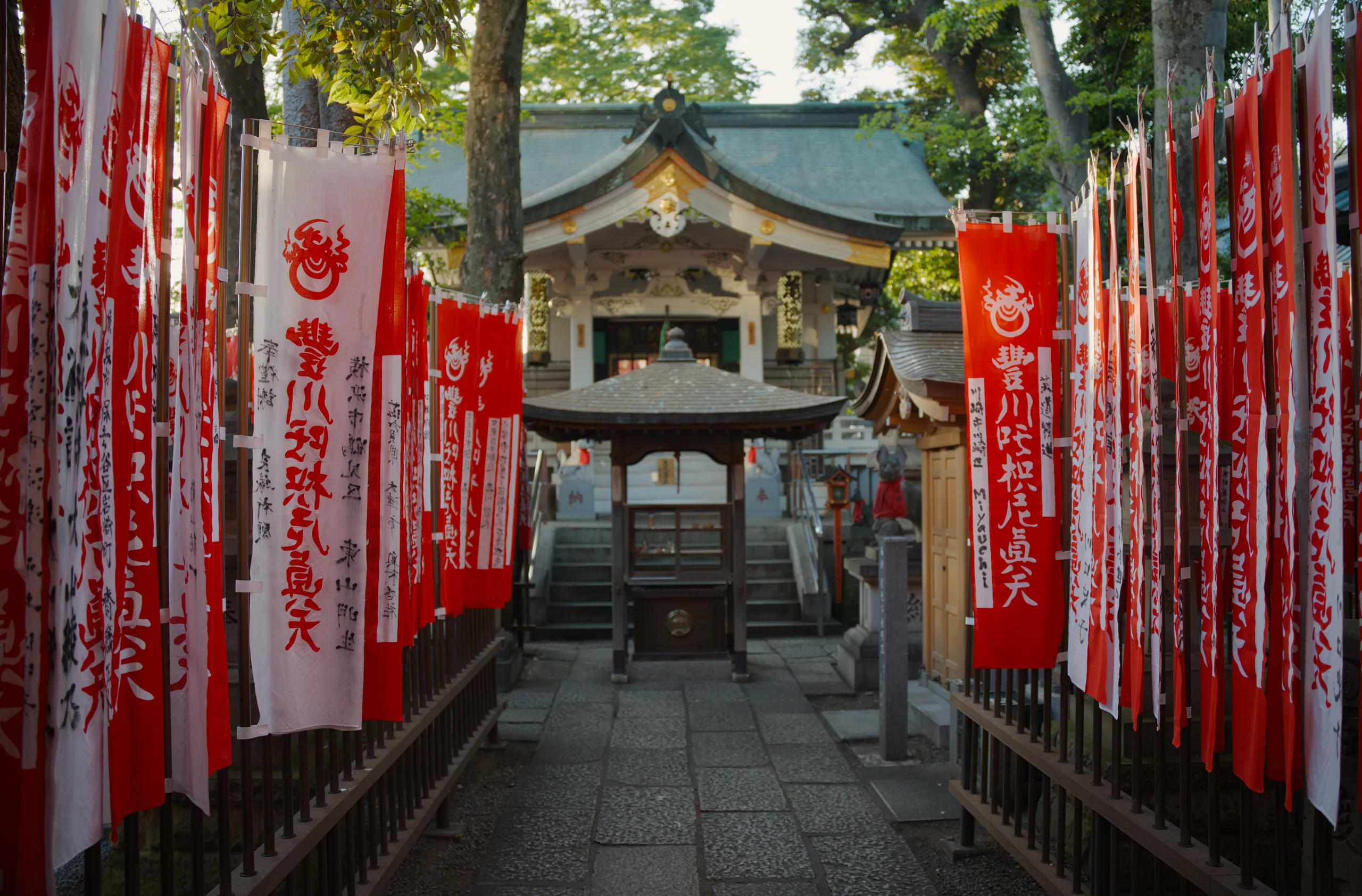 Red lanterns and banners line a covered shrine passageway with a wooden building visible beyond.